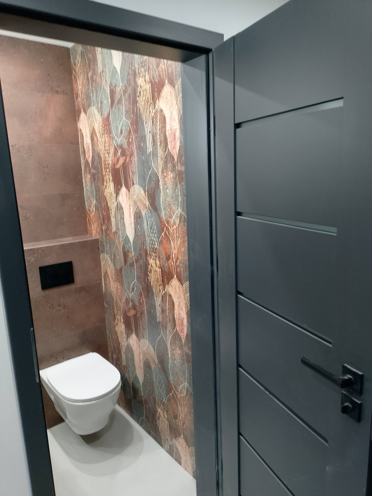 Wykończona toaleta w brązach z wzorzystą ścianą. Podwieszana miska WC, szara stolarka drzwiowa z poziomymi frezami. Nowoczesny design.
