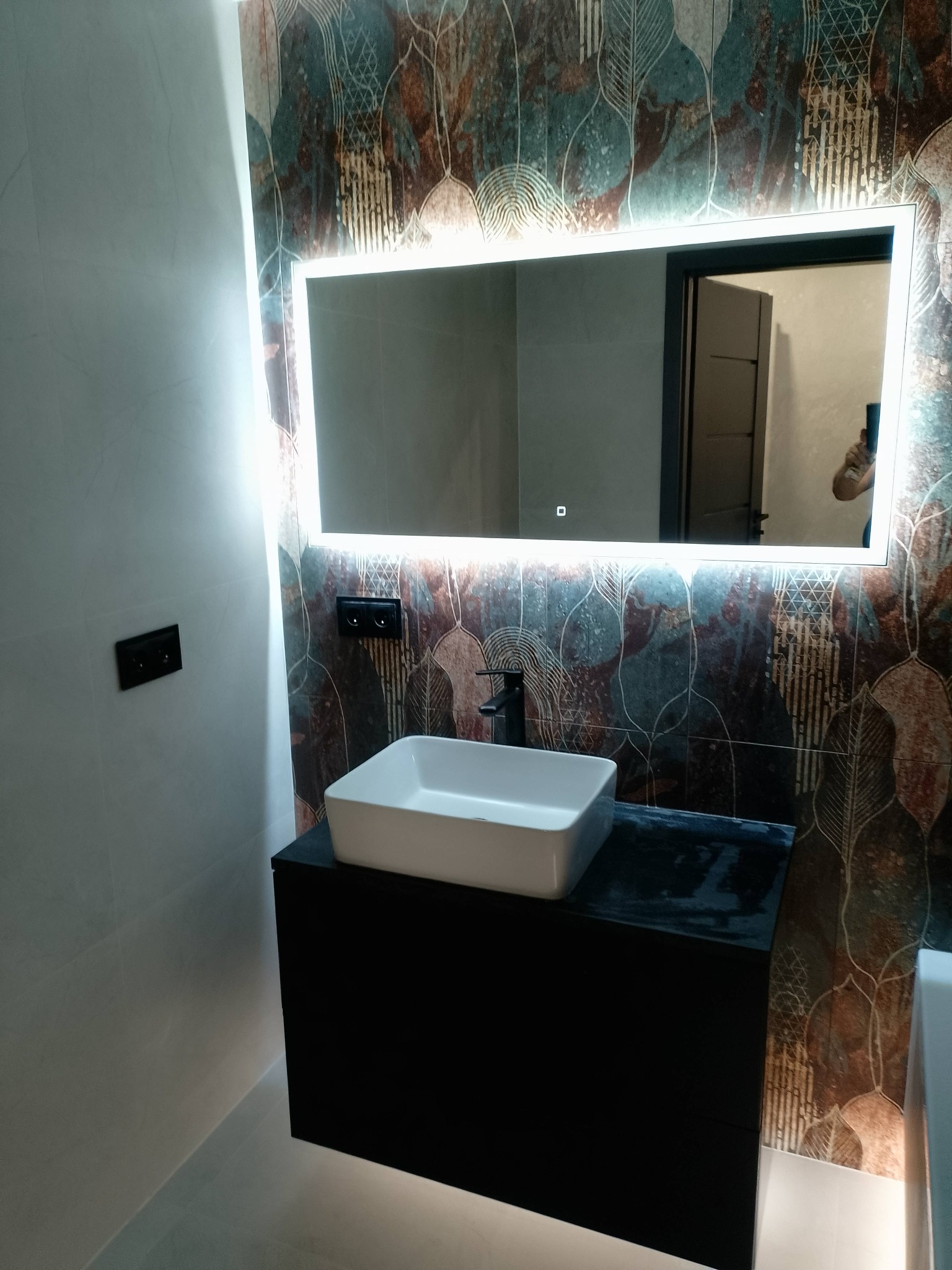 Nowoczesna łazienka z podświetlanym lustrem, białą umywalką na czarnej szafce i dekoracyjnymi płytkami w odcieniach brązu i turkusu. Widoczne czarne włączniki.