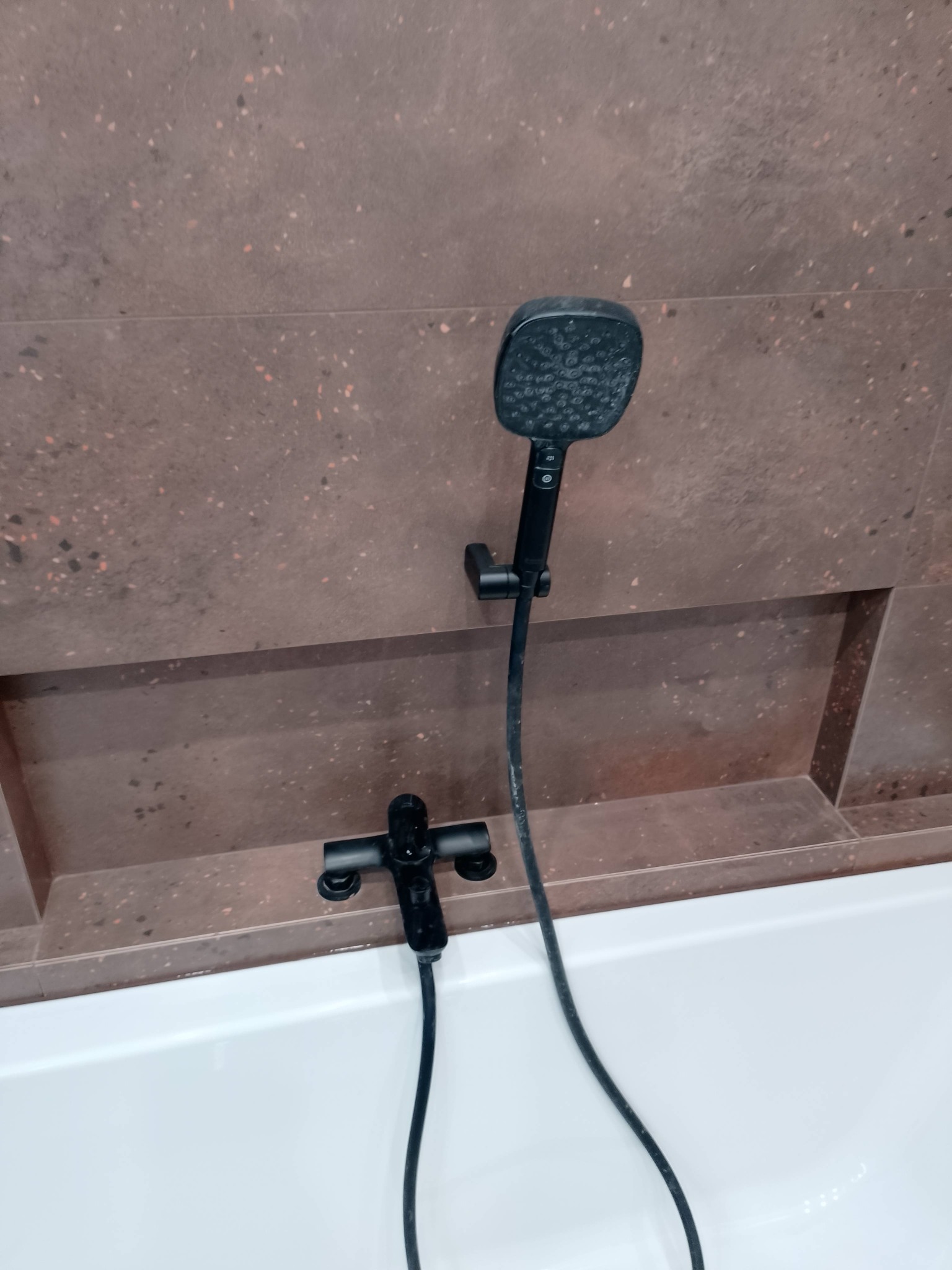 Nowoczesna, czarna armatura łazienkowa: bateria i słuchawka prysznicowa na tle brązowych płytek z wnęką nad białą wanną. Minimalistyczny design.