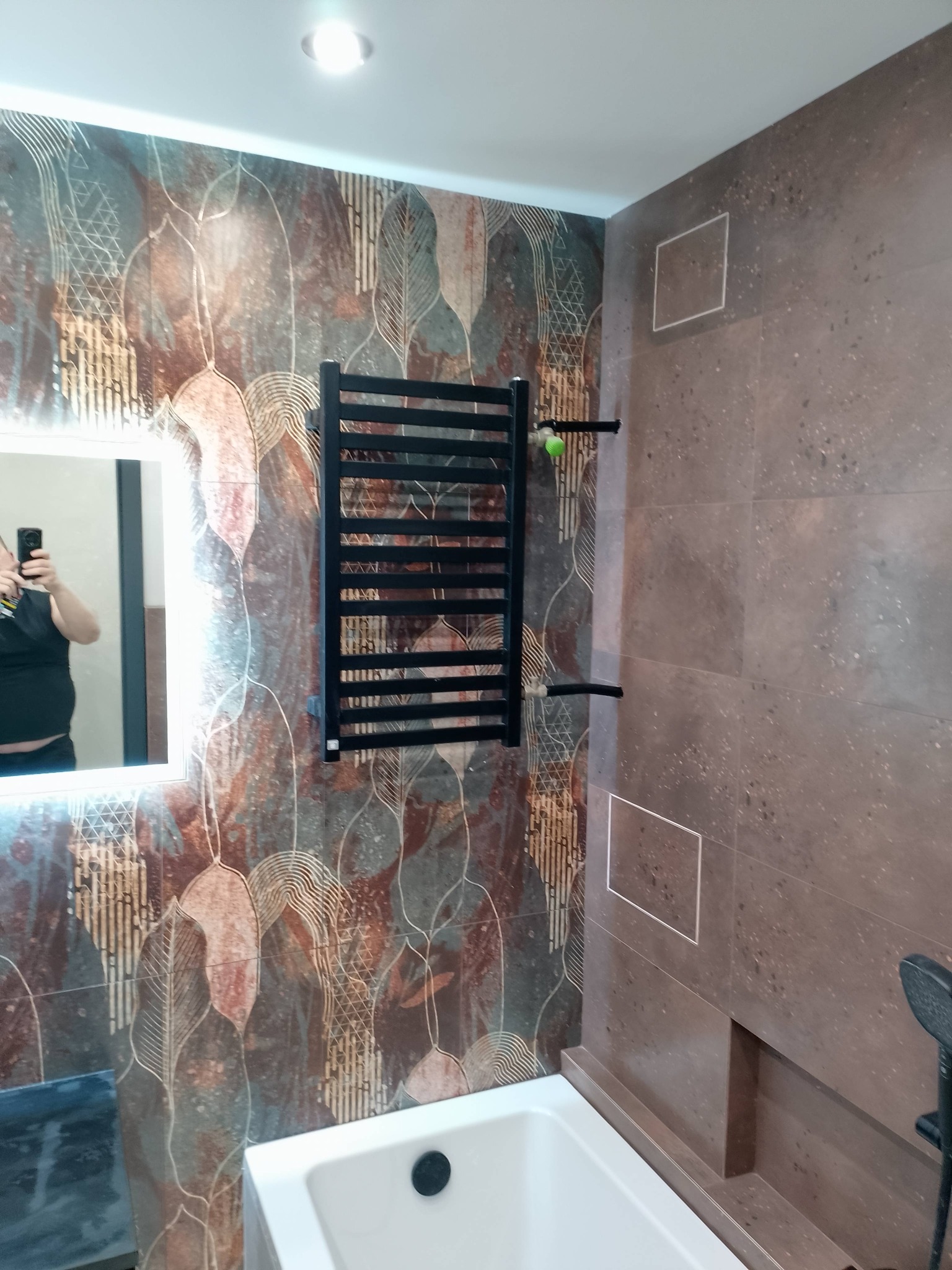 Wykończona łazienka z dekoracyjnymi płytkami w odcieniach brązu i beżu, czarnym grzejnikiem drabinkowym i podświetlanym lustrem. Widoczna wanna i fragment armatury.