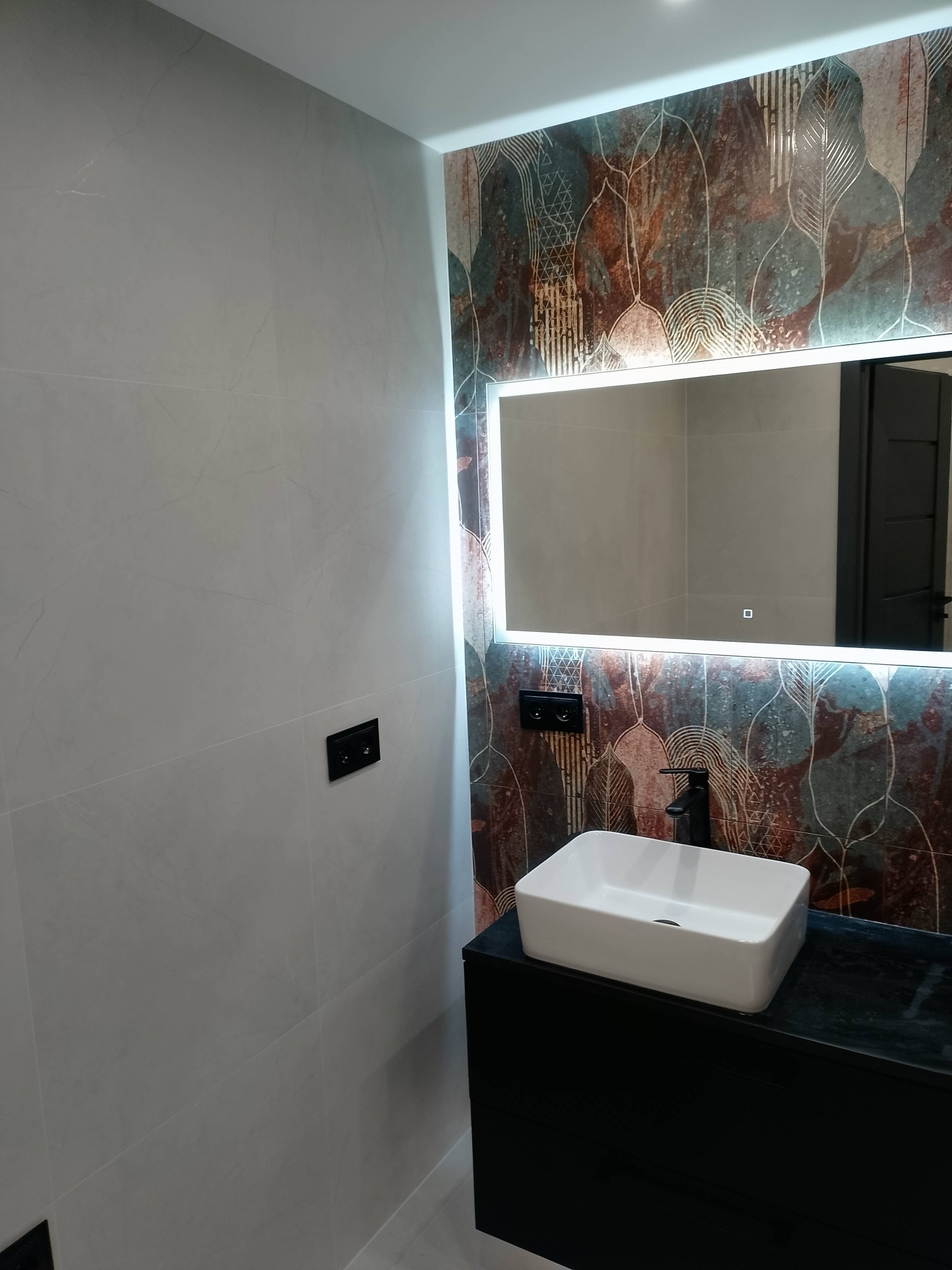 Nowoczesna łazienka z umywalką nablatową, podświetlanym lustrem i dekoracyjną ścianą w odcieniach brązu i turkusu. Minimalistyczny design, czarne akcenty.