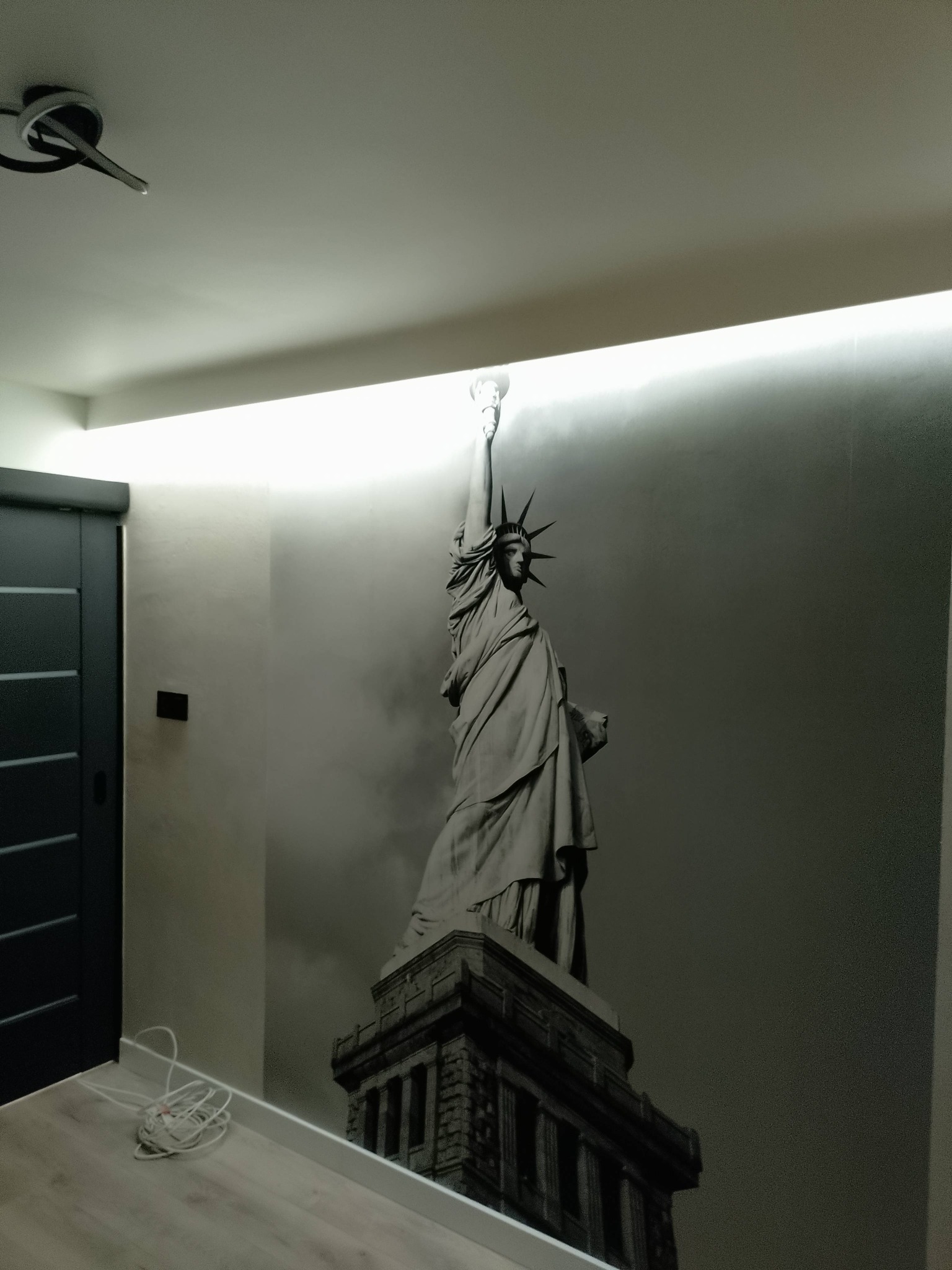 Nowoczesne wnętrze z fototapetą Statuy Wolności i oświetleniem LED. Ciemne drzwi przesuwne i jasna podłoga tworzą kontrast. Widoczne kable na podłodze.