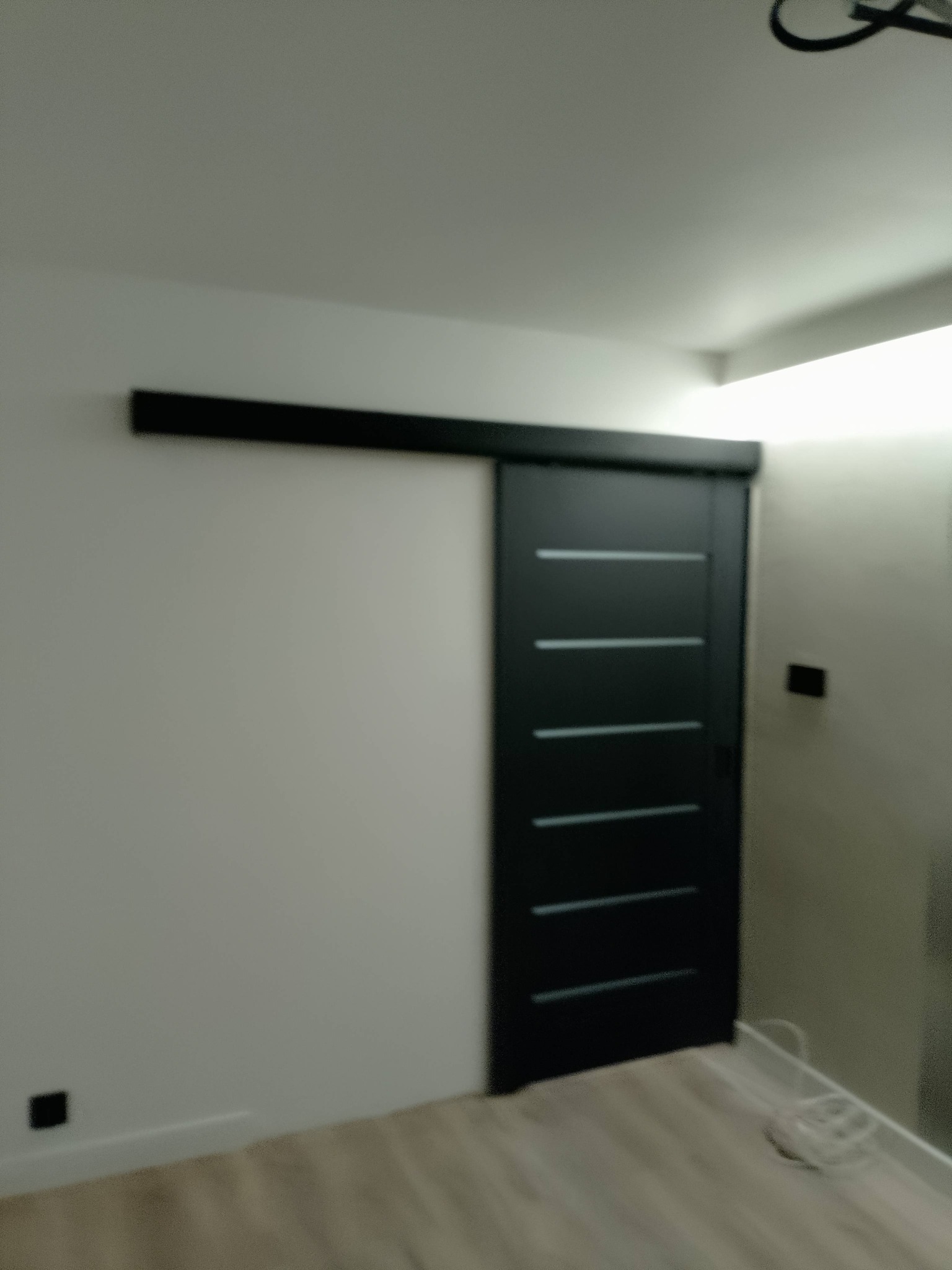 Nowoczesne, czarne drzwi przesuwne z poziomymi przeszkleniami, zamontowane w jasnym wnętrzu. Minimalistyczny design, widoczne gniazdko elektryczne na ścianie.