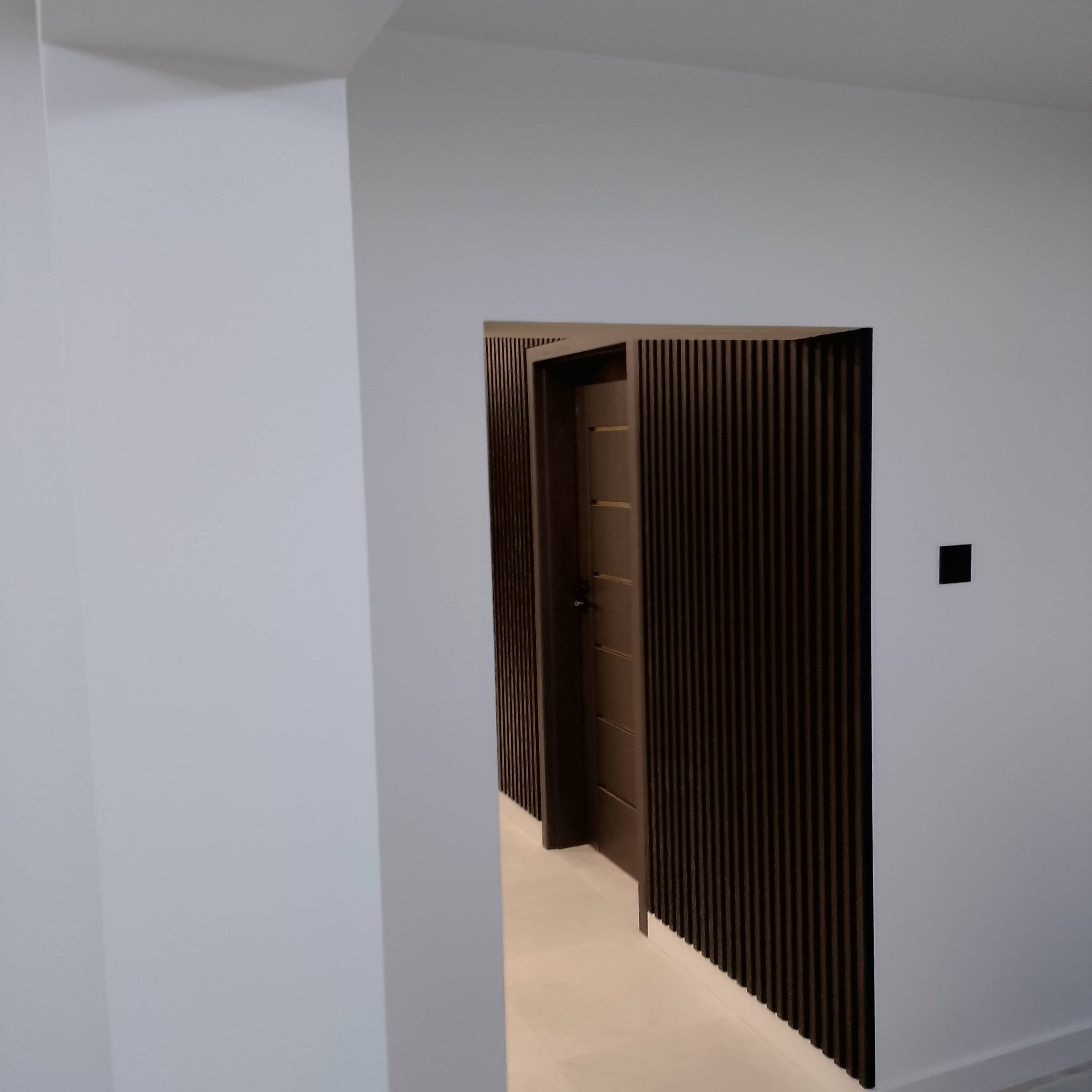Nowoczesne wykończenie wnętrza: ciemne lamele dekoracyjne okalają wejście do pokoju, kontrastując z jasnymi ścianami. Minimalistyczny design, czarny włącznik światła.