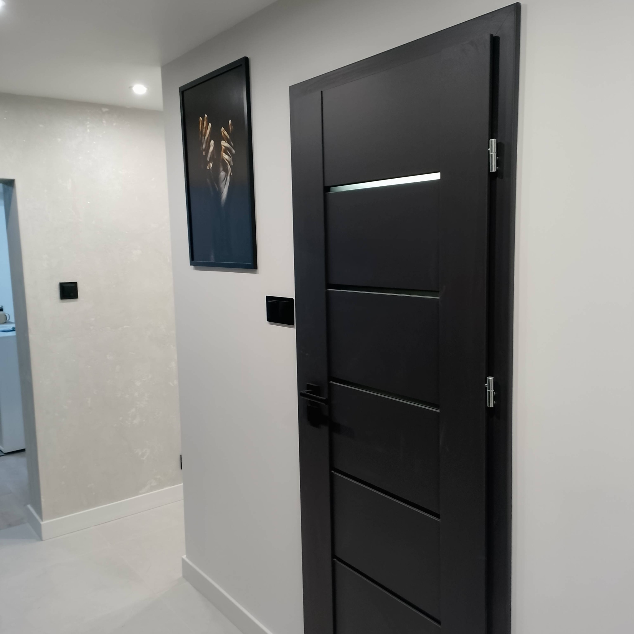 Ciemne, nowoczesne drzwi wewnętrzne z poziomymi frezowaniami i srebrnym akcentem, zamontowane w jasnym korytarzu z dekoracyjną ścianą i czarnym włącznikiem.