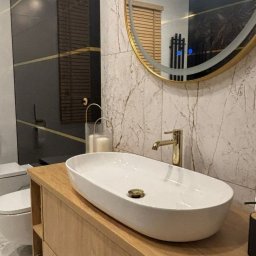 Firma remontowo-wykończeniowa PROFI-GIPS - Nowoczesna łazienka z owalną białą umywalką nablatową, złotą baterią i okrągłym lustrem w złotej ramie, marmurowe płytki na ścianie, czarna ściana z poziomymi złotymi paskami w tle.