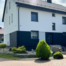 Firma remontowo-wykończeniowa PROFI-GIPS - Dwukondygnacyjny dom z białą elewacją na górze i ciemną na dole, z czarnymi oknami i dachem, otoczony zielenią i kostką brukową.