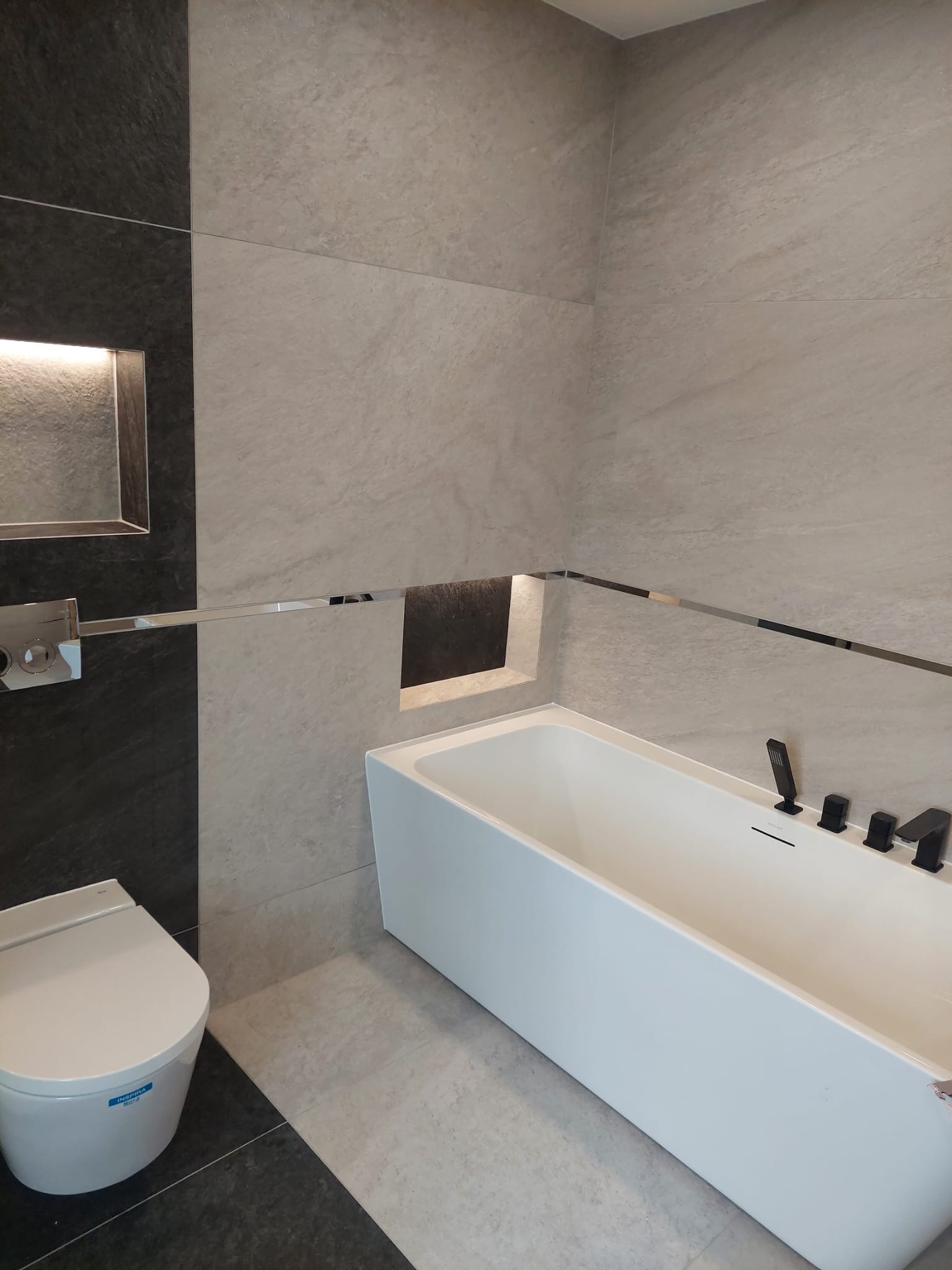 Nowoczesna łazienka z białą wanną, wiszącą toaletą i szarymi płytkami z oświetlonymi niszami.