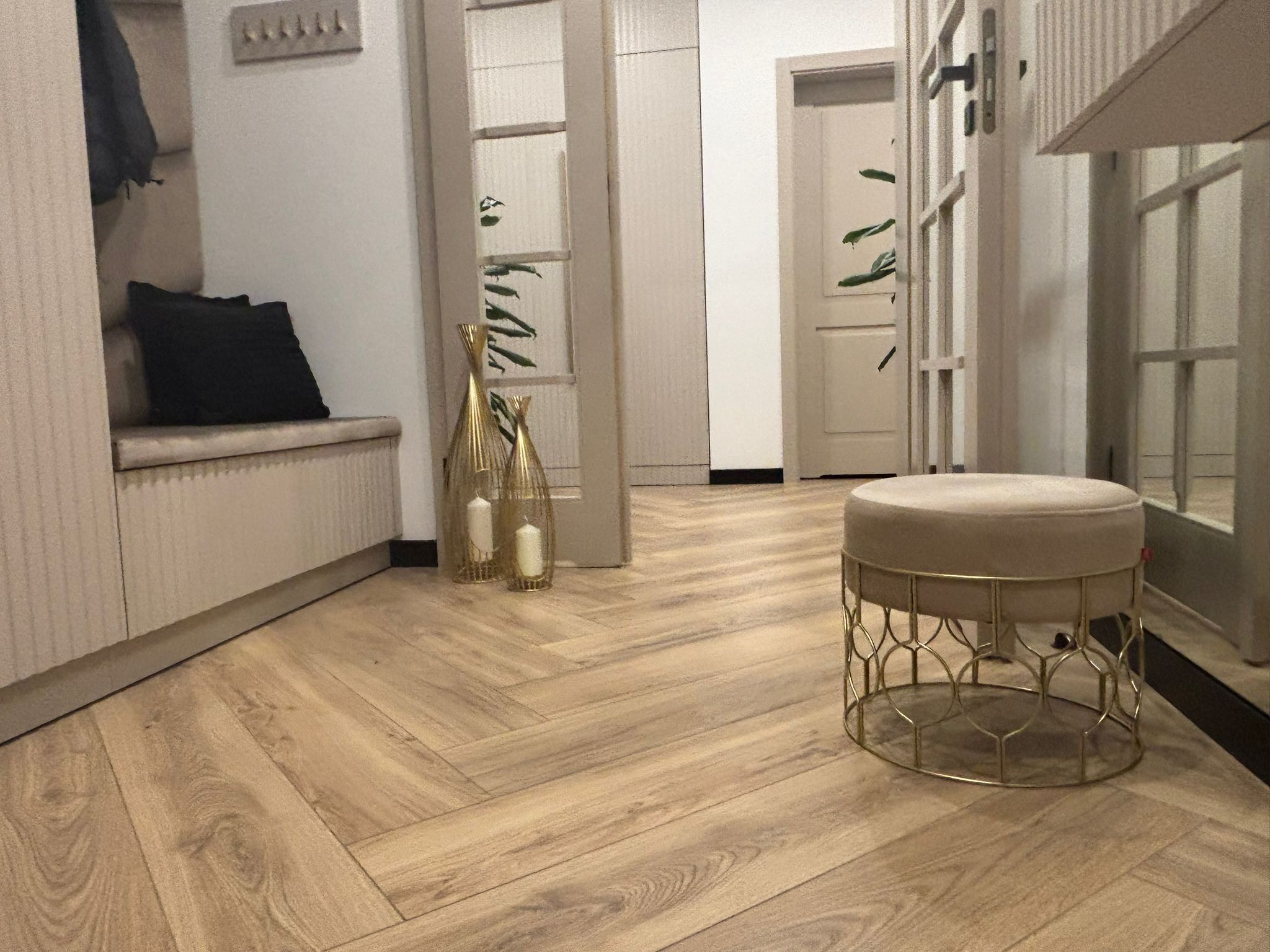 Beżowy przedpokój z parkietem ułożonym w jodełkę, dekoracyjnymi złotymi wazonami i pufą na metalowej podstawie.