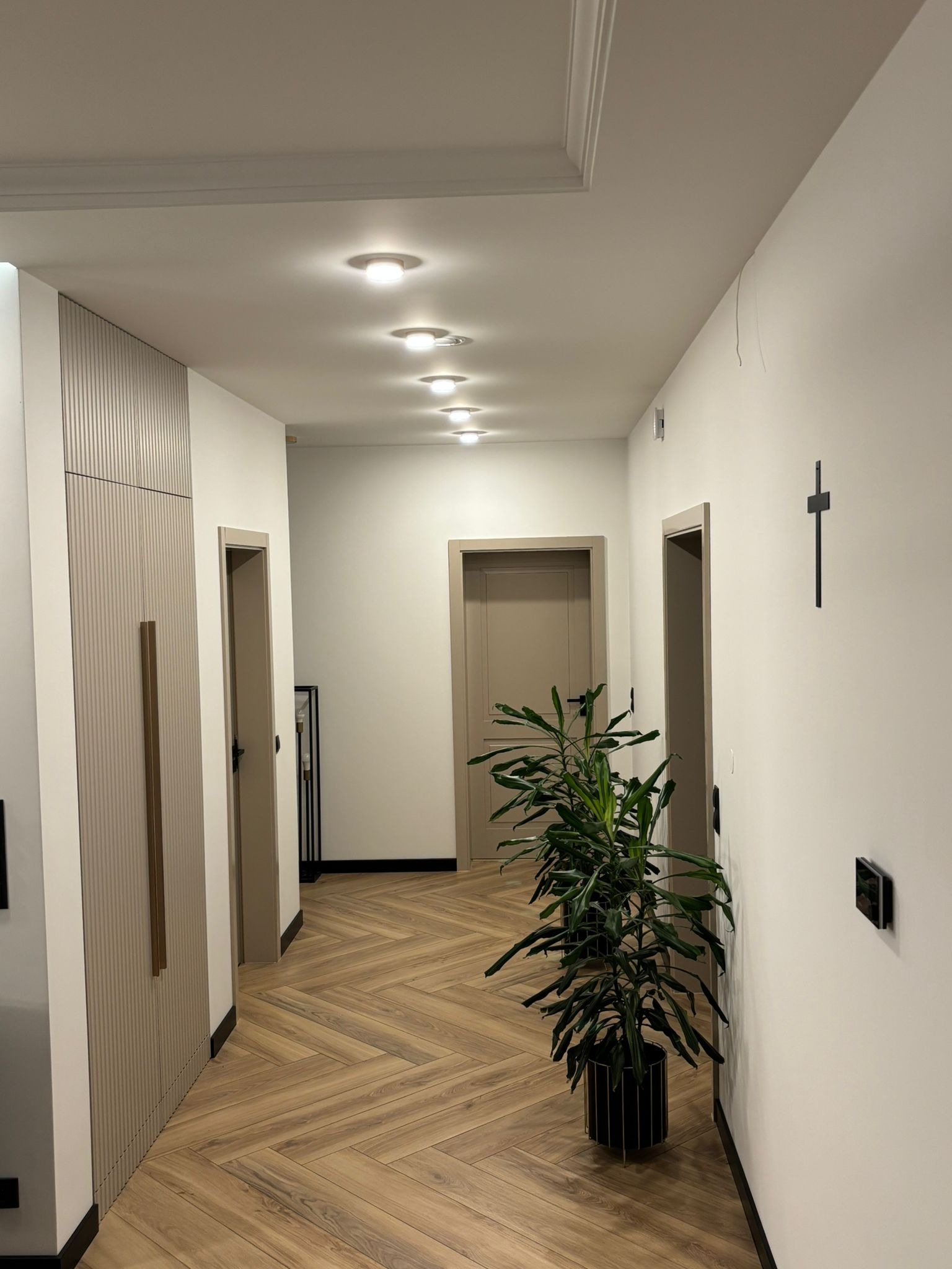 Nowoczesny korytarz z parkietem w jodełkę, białymi ścianami, beżowymi drzwiami, wbudowaną szafą z pionowymi paskami, rzędem sufitowych lamp i dekoracyjnym krzyżem na ścianie.