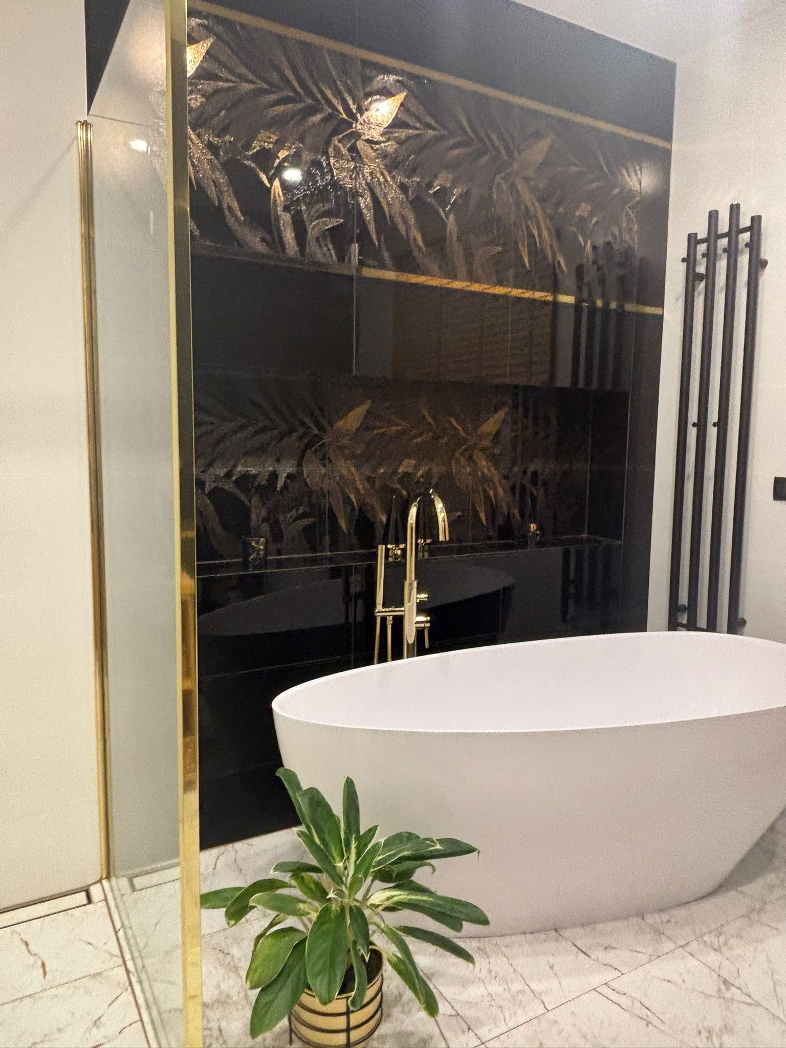 Nowoczesna łazienka z czarnymi płytkami z motywem liści, wolnostojącą białą wanną, złotą armaturą i czarnym grzejnikiem drabinkowym.