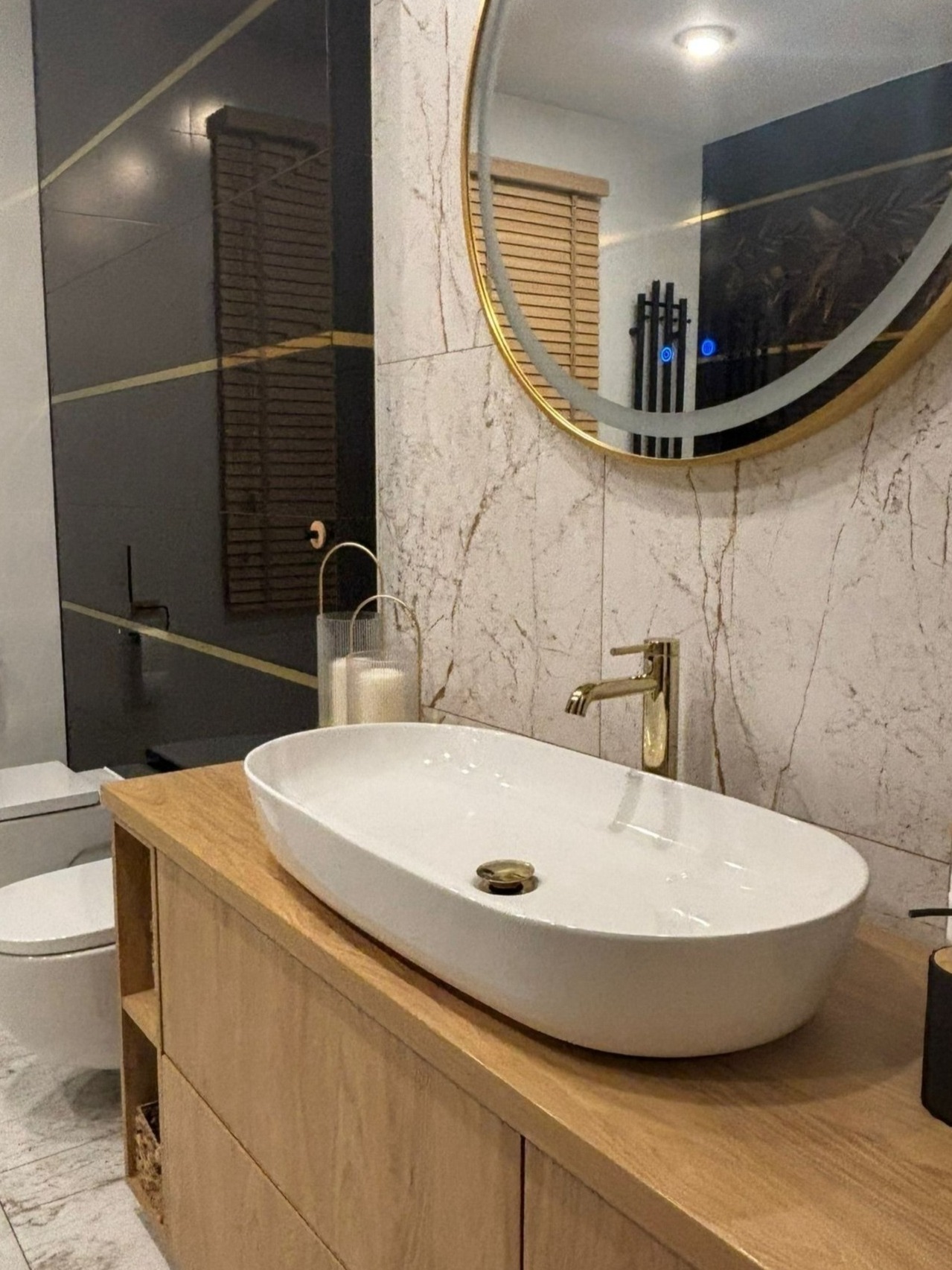 Nowoczesna łazienka z owalną białą umywalką nablatową, złotą baterią i okrągłym lustrem w złotej ramie, marmurowe płytki na ścianie, czarna ściana z poziomymi złotymi paskami w tle.