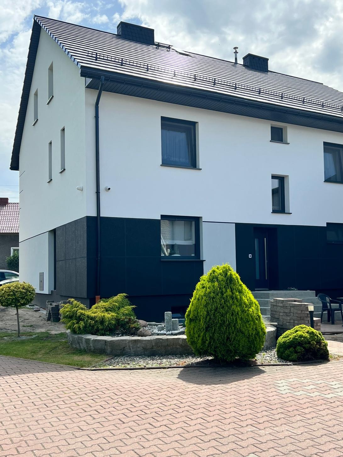 Dwukondygnacyjny dom z białą elewacją na górze i ciemną na dole, z czarnymi oknami i dachem, otoczony zielenią i kostką brukową.