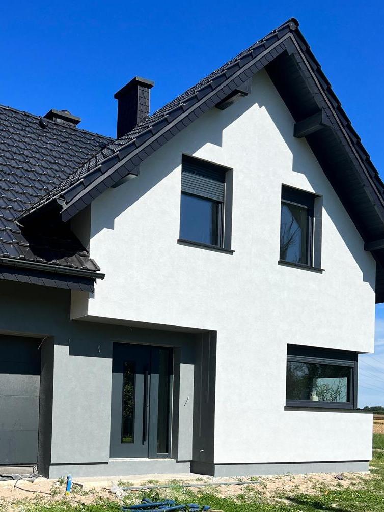 Nowoczesny dom jednorodzinny z białą elewacją o fakturze baranka, grafitową stolarką okienną i drzwiami, oraz ciemnoszarą dachówką, na tle błękitnego nieba.