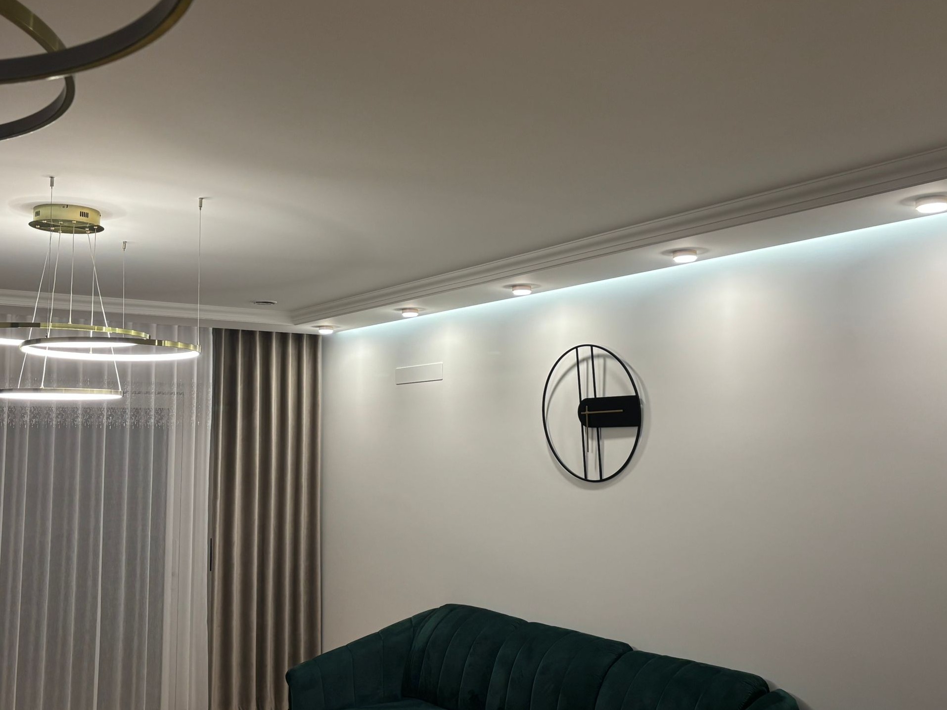 Nowoczesny salon z szarą ścianą, okrągłym zegarem, listwą oświetleniową pod sufitem i designerską lampą wiszącą.