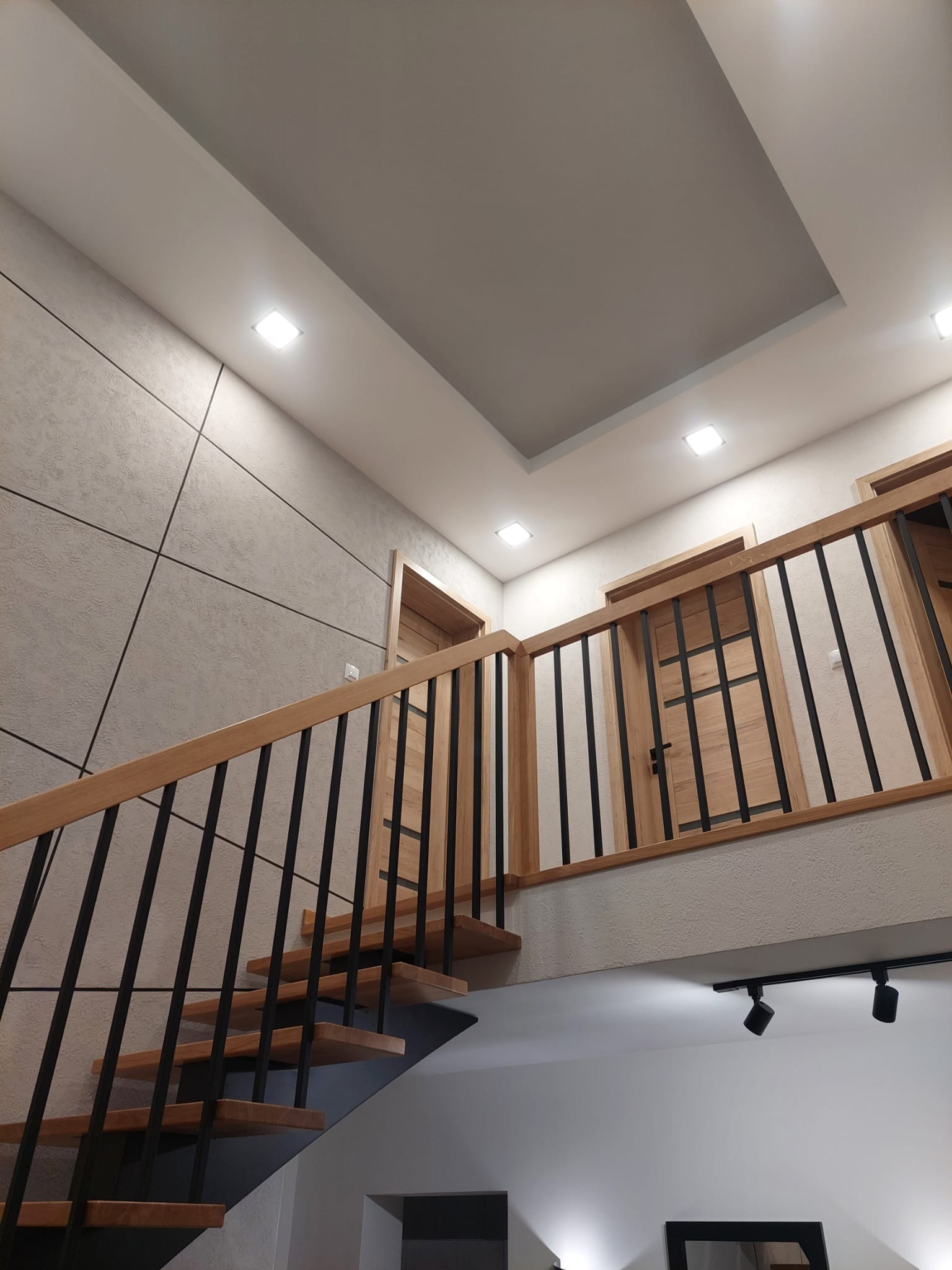 Nowoczesna klatka schodowa z drewnianymi schodami i czarną balustradą, oświetlona punktowymi lampami, z widoczną ścianą wykończoną dekoracyjnym tynkiem i drewnianymi drzwiami z czarnymi akcentami.