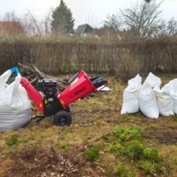 Czerwona rozdrabniarka do gałęzi marki Cedrus z pełnym workiem zrębków i pięcioma workami gotowymi do transportu na tle żywopłotu i zabudowań.