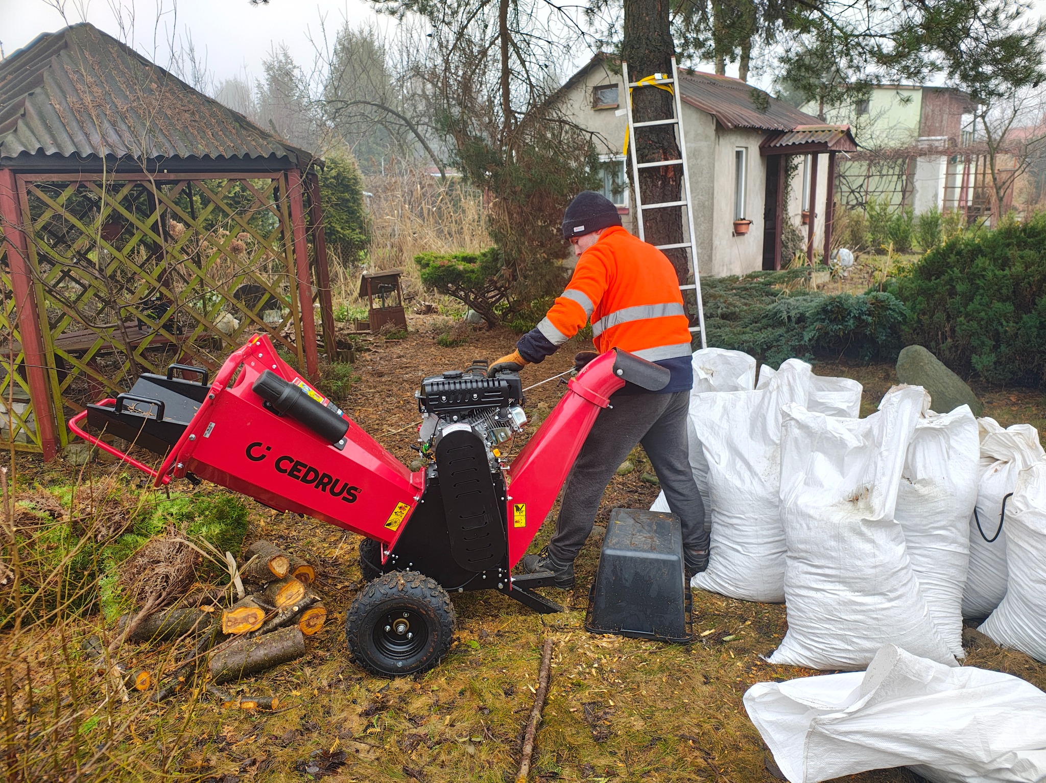 Pracownik w odblaskowej kurtce obsługuje czerwoną rębarkę Cedrus, obok sterta pociętych gałęzi i worki zrębków, w tle ogród z altaną i domem.