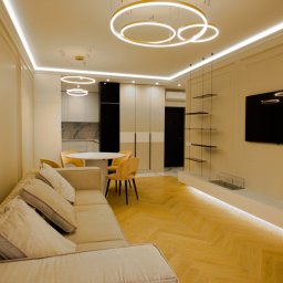 Kultura wnętrza - Salon z aneksem kuchennym w odcieniach beżu i bieli, z parkietem w jodełkę, oświetleniem LED, okrągłym stołem, żółtymi krzesłami, szklanymi półkami i telewizorem na ścianie z dekoracyjnymi listwami.