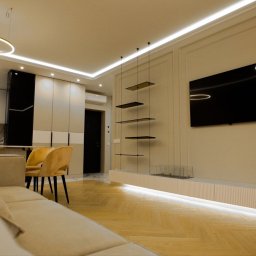 Kultura wnętrza - Nowoczesny salon z parkietem w jodełkę, minimalistyczną szafką RTV z podświetleniem LED, wiszącymi półkami na linkach i telewizorem na ścianie w kremowych panelach.