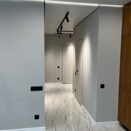 Kultura wnętrza - Nowoczesny korytarz z szarymi ścianami, białymi płytkami imitującymi marmur z brązowymi żyłami, czarnymi włącznikami i gniazdkami, oraz minimalistycznym oświetleniem sufitowym.