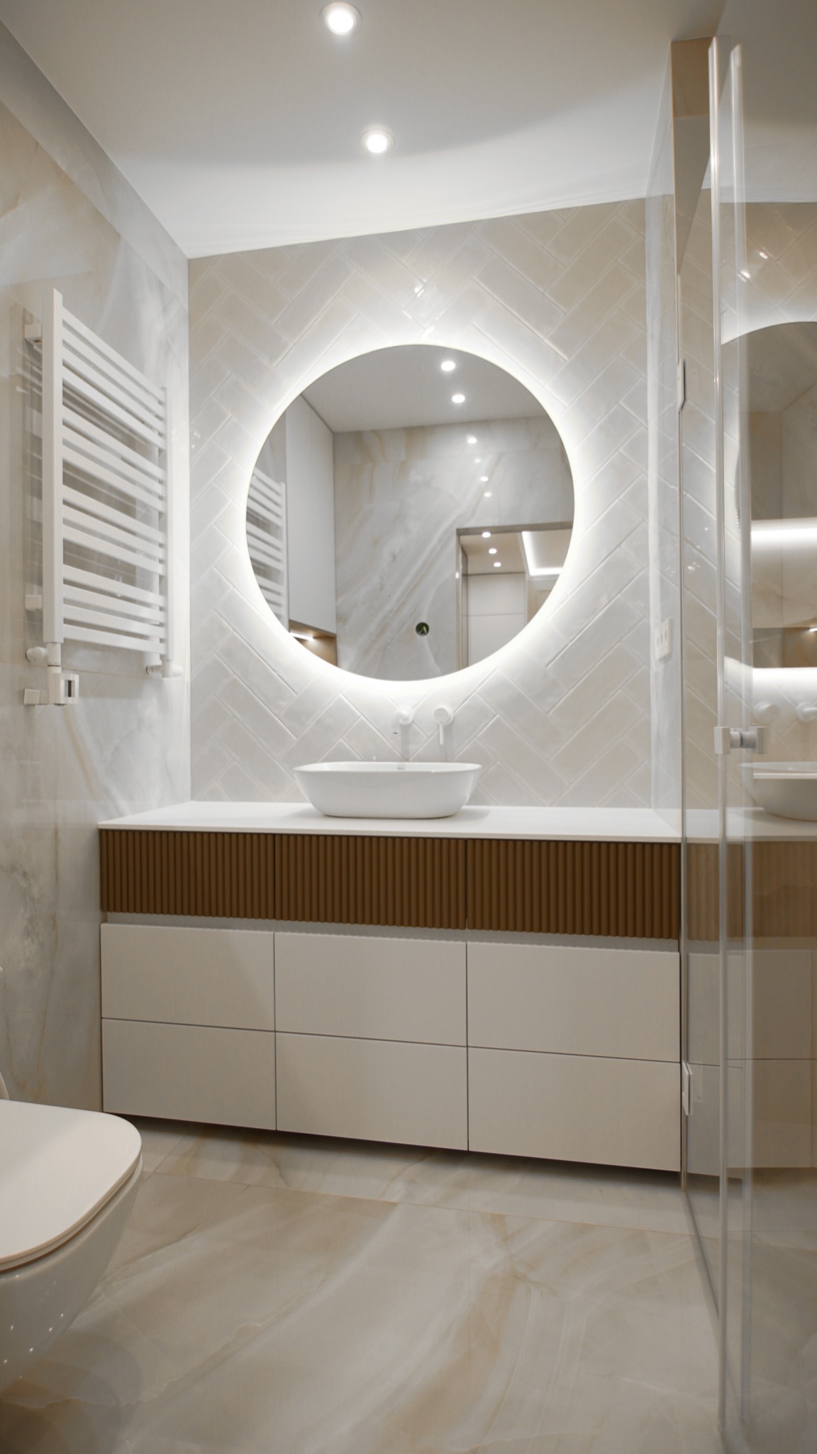 Nowoczesna łazienka z okrągłym lustrem podświetlanym LED, umywalką nablatową, białymi szafkami z drewnianymi akcentami i grzejnikiem drabinkowym.