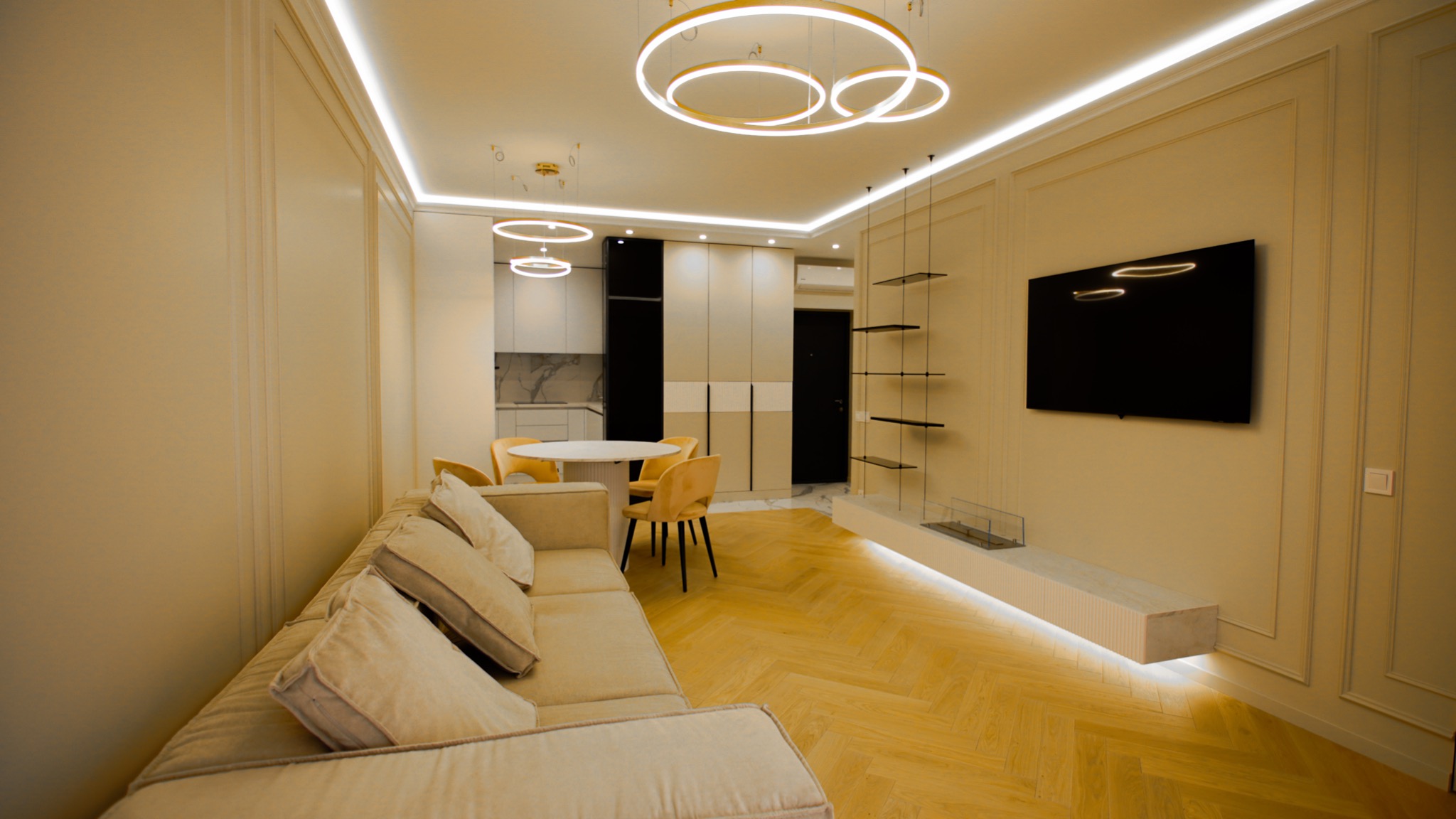 Salon z aneksem kuchennym w odcieniach beżu i bieli, z parkietem w jodełkę, oświetleniem LED, okrągłym stołem, żółtymi krzesłami, szklanymi półkami i telewizorem na ścianie z dekoracyjnymi listwami.