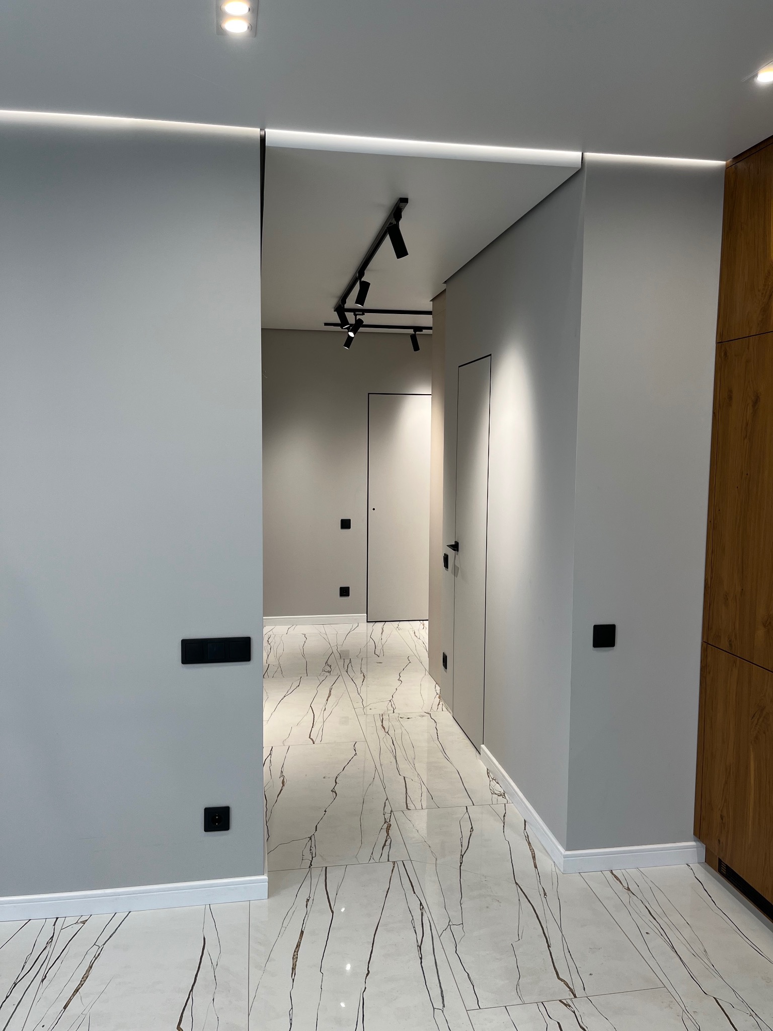 Nowoczesny korytarz z szarymi ścianami, białymi płytkami imitującymi marmur z brązowymi żyłami, czarnymi włącznikami i gniazdkami, oraz minimalistycznym oświetleniem sufitowym.