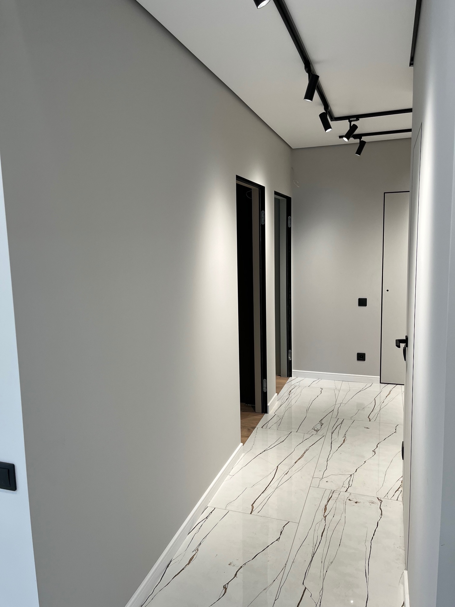 Nowoczesny korytarz z szarymi ścianami, białą podłogą z marmurowym wzorem, czarnymi drzwiami i oświetleniem szynowym