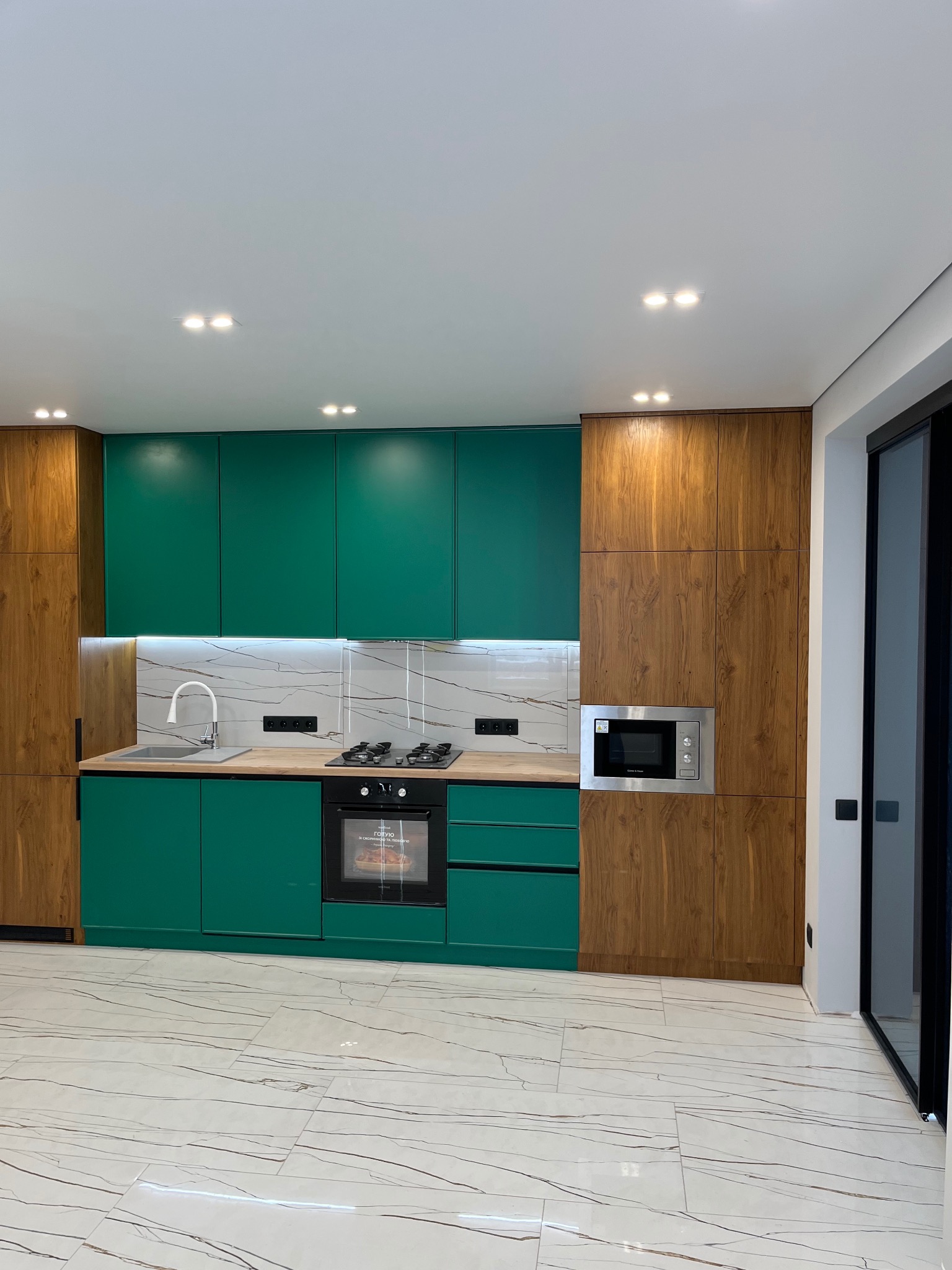 Nowoczesna kuchnia z zielonymi szafkami i drewnianymi elementami, marmurowym backsplashiem, czarnym piekarnikiem i płytą grzewczą, oświetlona punktowymi lampami sufitowymi, na podłodze białe płytki...