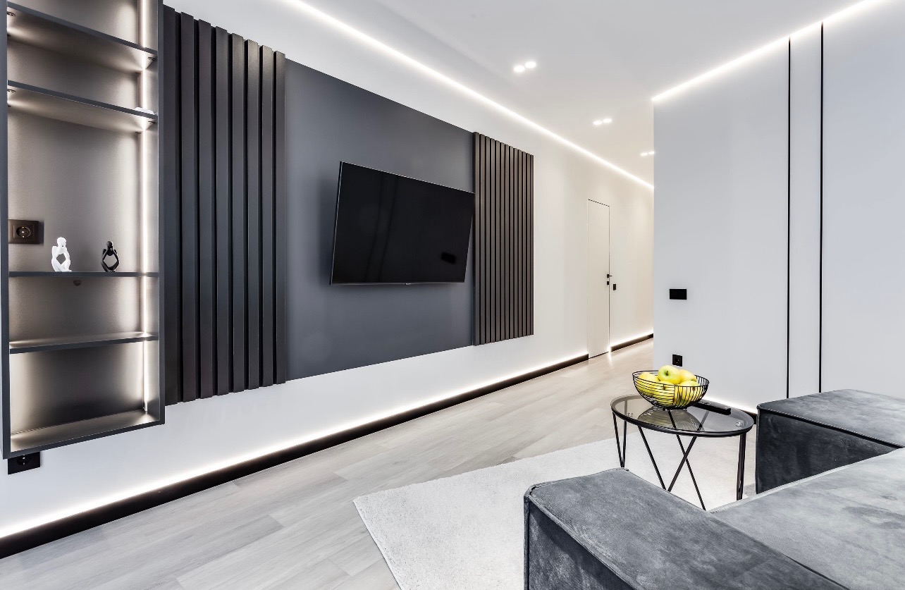Minimalistyczny salon z szarą ścianą akcentową, panele dekoracyjne, podświetlenie LED, wiszący telewizor i szklany stolik kawowy z owocami.