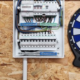 Krzysztof Michalik - Otwarta skrzynka rozdzielcza z licznikiem energii elektrycznej i zabezpieczeniami, zamontowana na ścianie z płyty OSB, obok tarczy do darta.