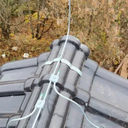 Widok z góry na fragment dachu pokrytego czarną dachówką z zamontowaną instalacją odgromową, zabezpieczoną metalowymi uchwytami i złączkami, na tle jesiennych drzew i zabudowań.