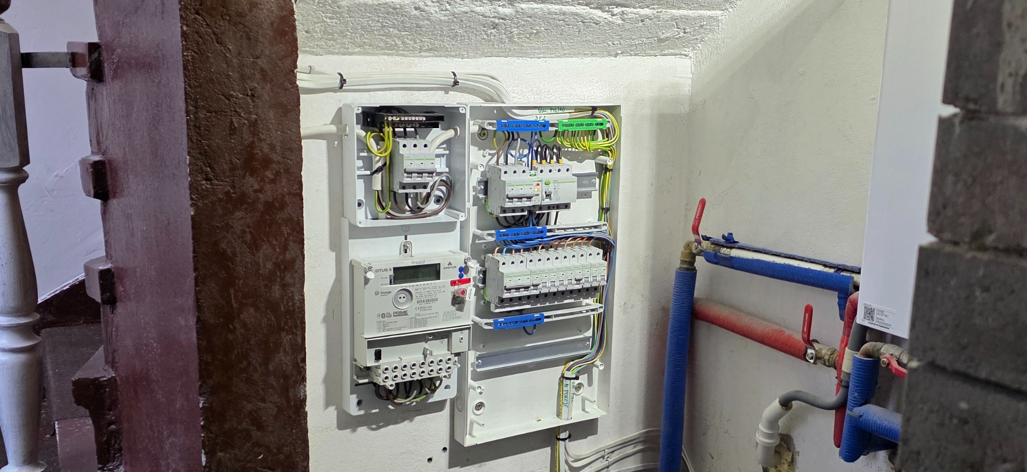 Otwarta skrzynka z bezpiecznikami i licznikiem energii elektrycznej zamontowana na ścianie w korytarzu obok rur centralnego ogrzewania i schodów z drewnianą balustradą.