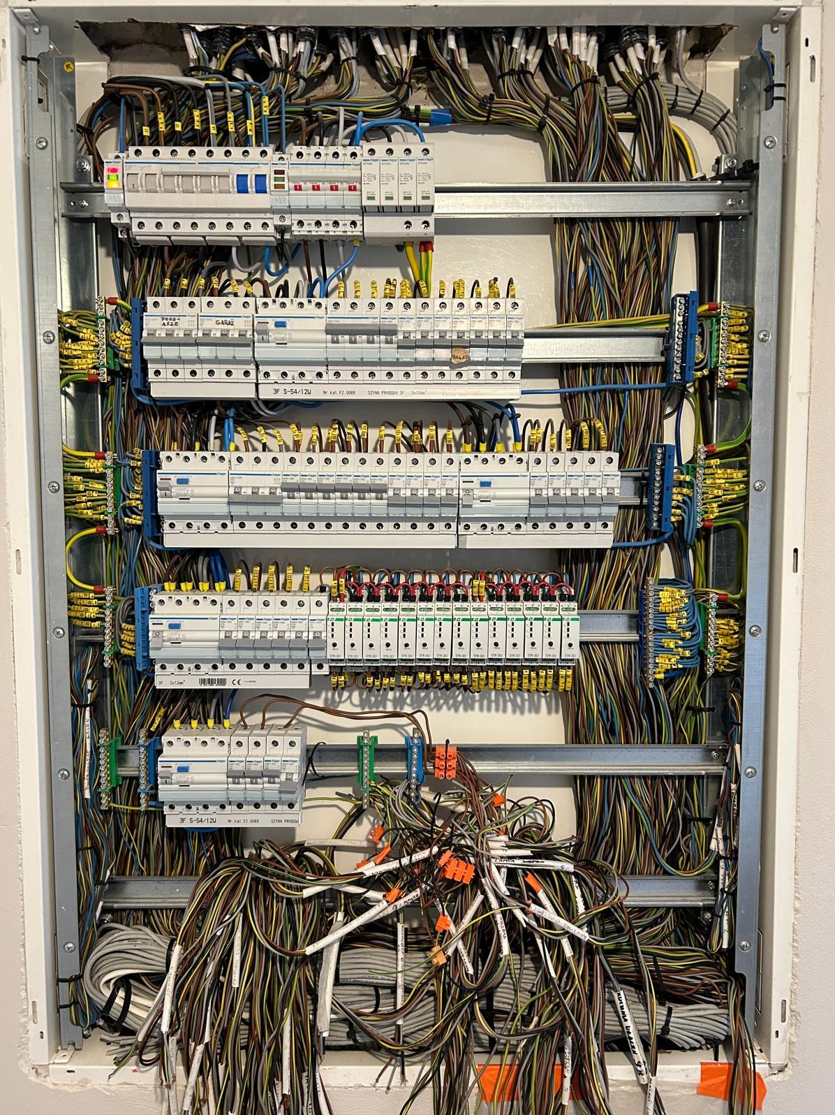 Otwarta skrzynka elektryczna z licznymi bezpiecznikami i przewodami o różnych kolorach, częściowo uporządkowanymi, a częściowo tworzącymi nieuporządkowany gąszcz na dole, widoczne oznaczenia...