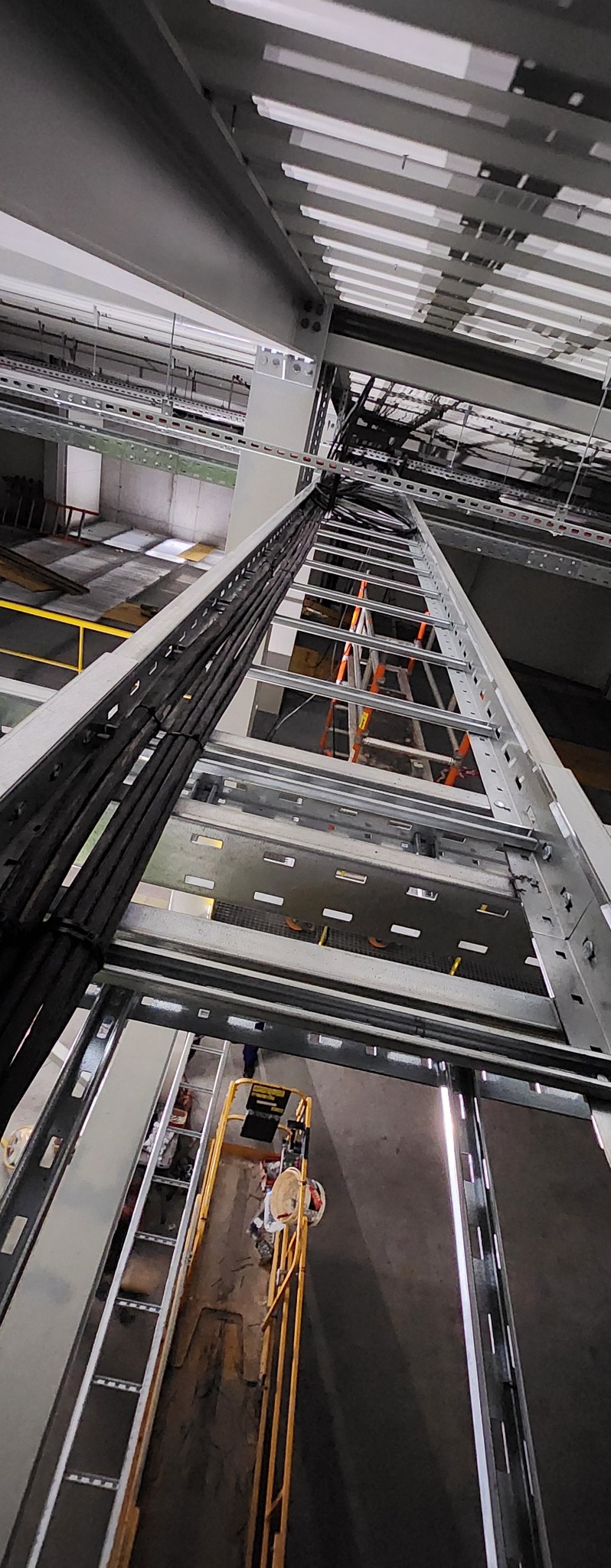 Montaż tras kablowych w budynku przemysłowym, widok z dołu na drabinki kablowe z ułożonymi wiązkami przewodów, pracownik na podnośniku koszowym w tle.
