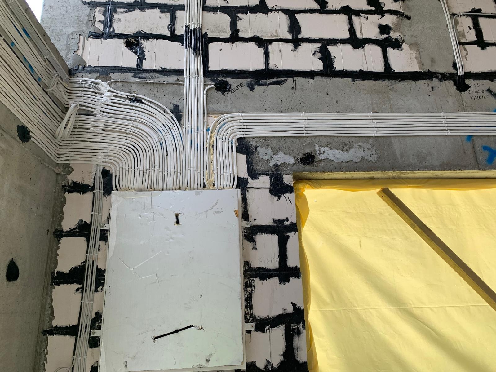 Układanie białych kabli elektrycznych w korytach na ścianie z białej cegły, zabezpieczone żółtą folią okno, widoczna skrzynka elektryczna.