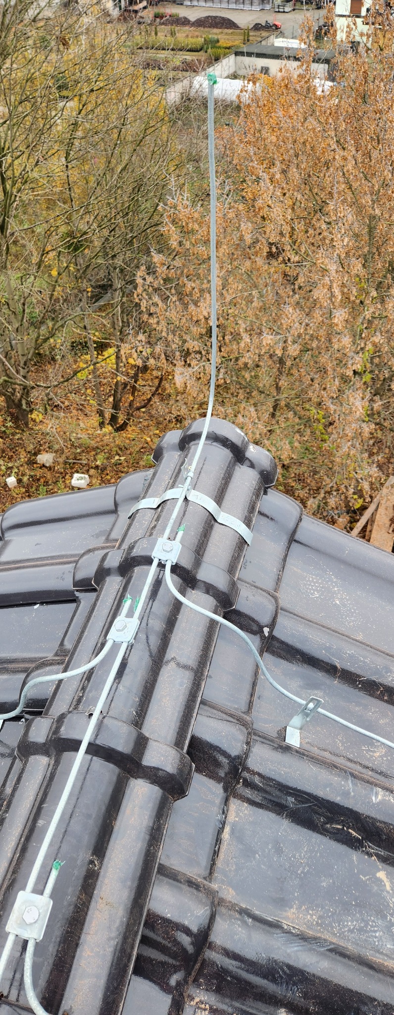 Widok z góry na fragment dachu pokrytego czarną dachówką z zamontowaną instalacją odgromową, zabezpieczoną metalowymi uchwytami i złączkami, na tle jesiennych drzew i zabudowań.