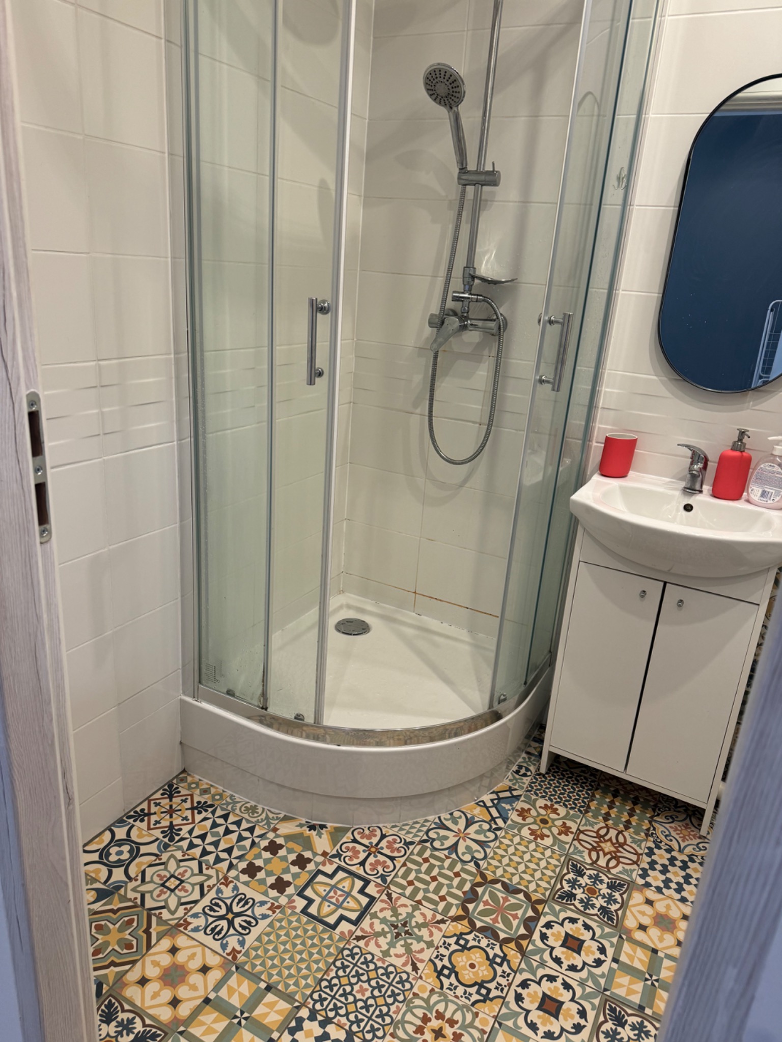 Wykończona łazienka w Ptaszkowej z kabiną prysznicową, umywalką z szafką i wzorzystą podłogą z płytek. Widoczne detale to owalne lustro w ciemnej ramie i czerwone akcenty w postaci kubka...