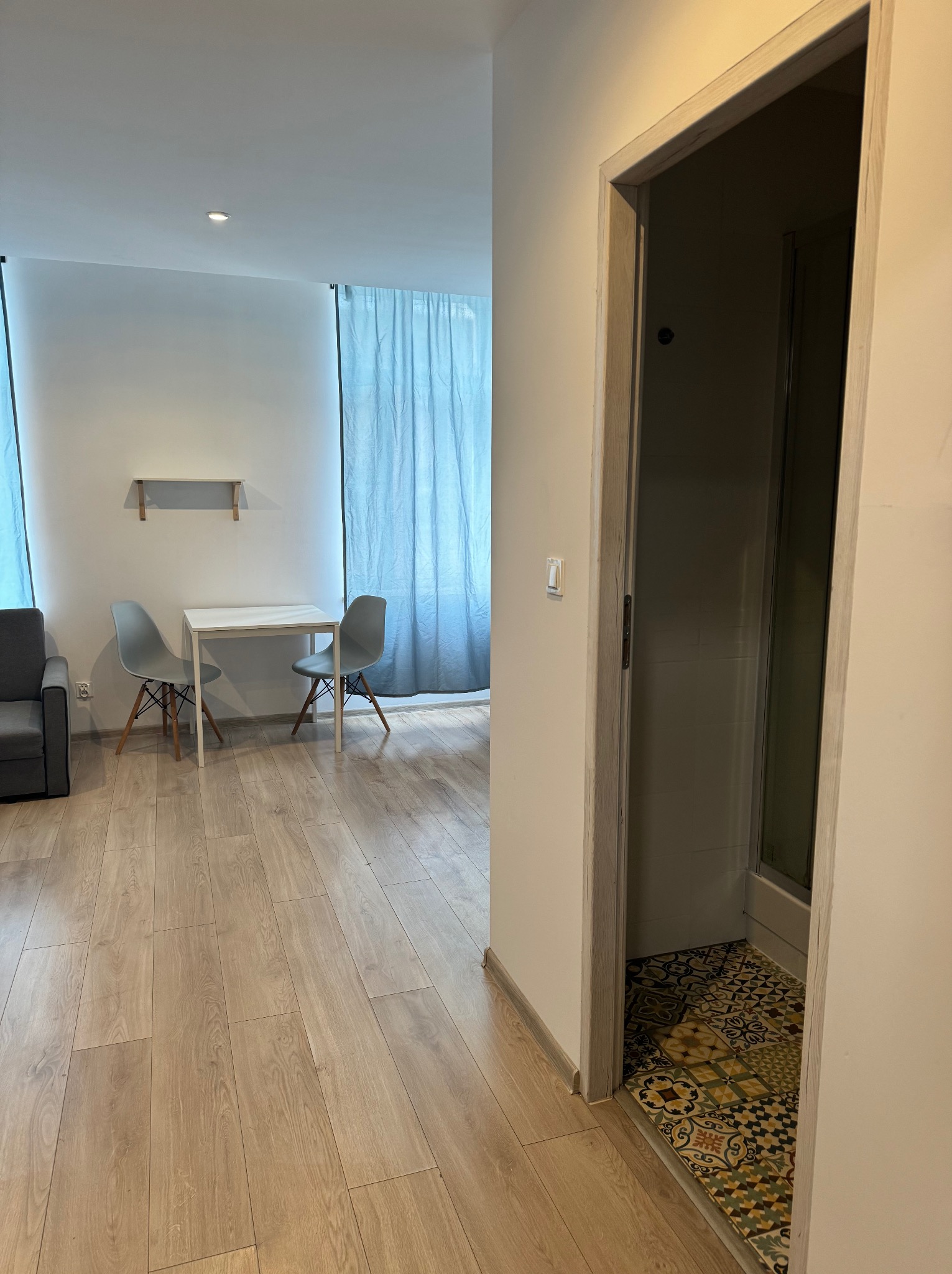 Wnętrze mieszkania po remoncie: parkiet z paneli, widok na łazienkę z wzorzystą podłogą, stół z krzesłami i zasłony w tle.