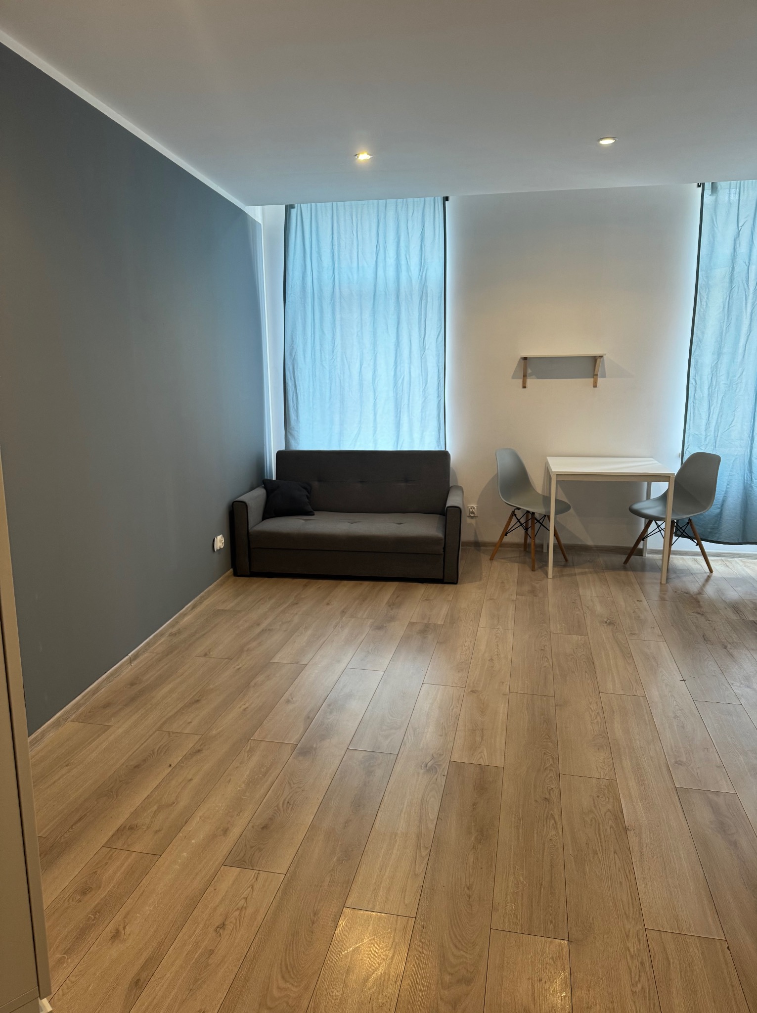 Wnętrze pokoju po remoncie: szara ściana kontrastuje z jasną podłogą z paneli, minimalistyczna sofa z czarną poduszką, stół z dwoma krzesłami i błękitne zasłony.