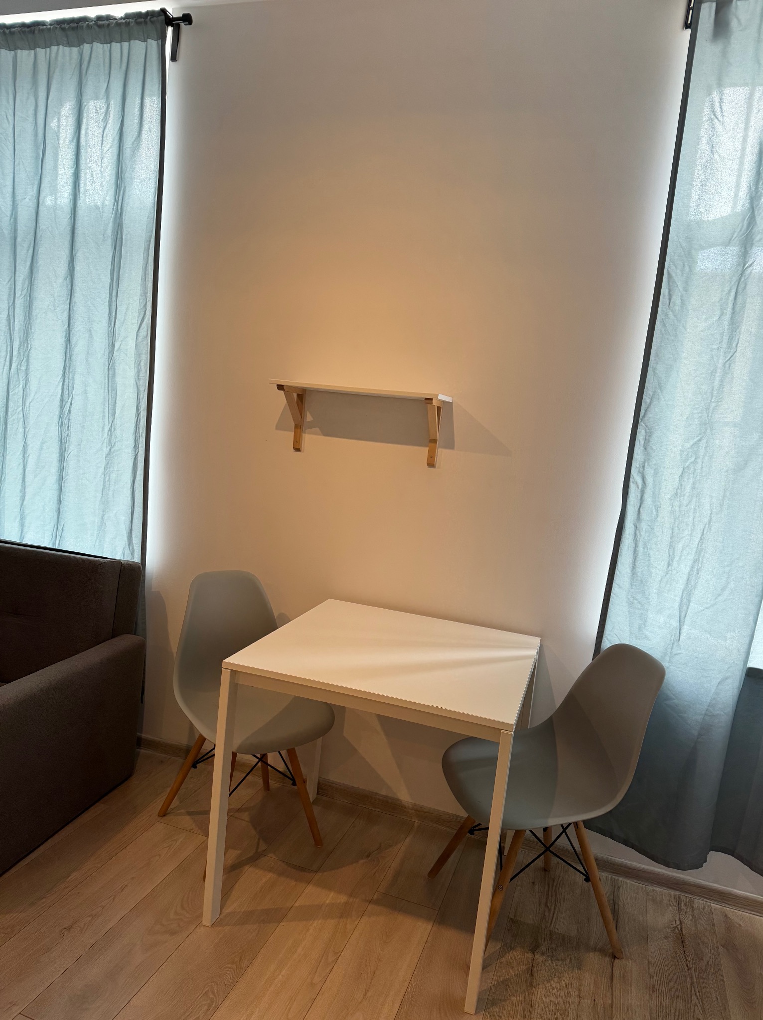 Jasne wnętrze pokoju z białym stołem i dwoma szarymi krzesłami, na ścianie zamontowana mała półka z jasnego drewna, zasłony w odcieniach błękitu w oknach.