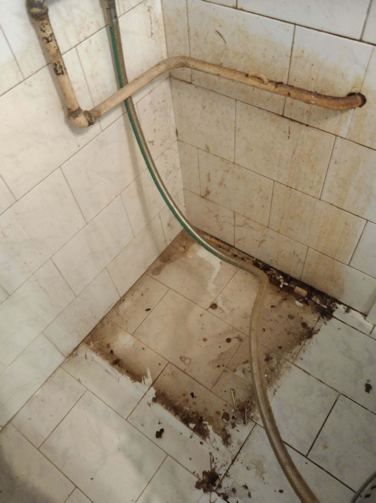 Zanieczyszczona podłoga i ściany wyłożone białymi płytkami w rogu pomieszczenia, widoczne stare rury i węże, resztki gruzu i brudu na fugach.