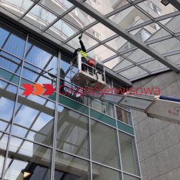Grupa Serwisowa sp z o.o. - Pracownik w podnośniku czyści szklany dach budynku biurowego. Widoczne odbicia w oknach i konstrukcja dachu. Ujęcie z dołu, z perspektywy przechodnia.
