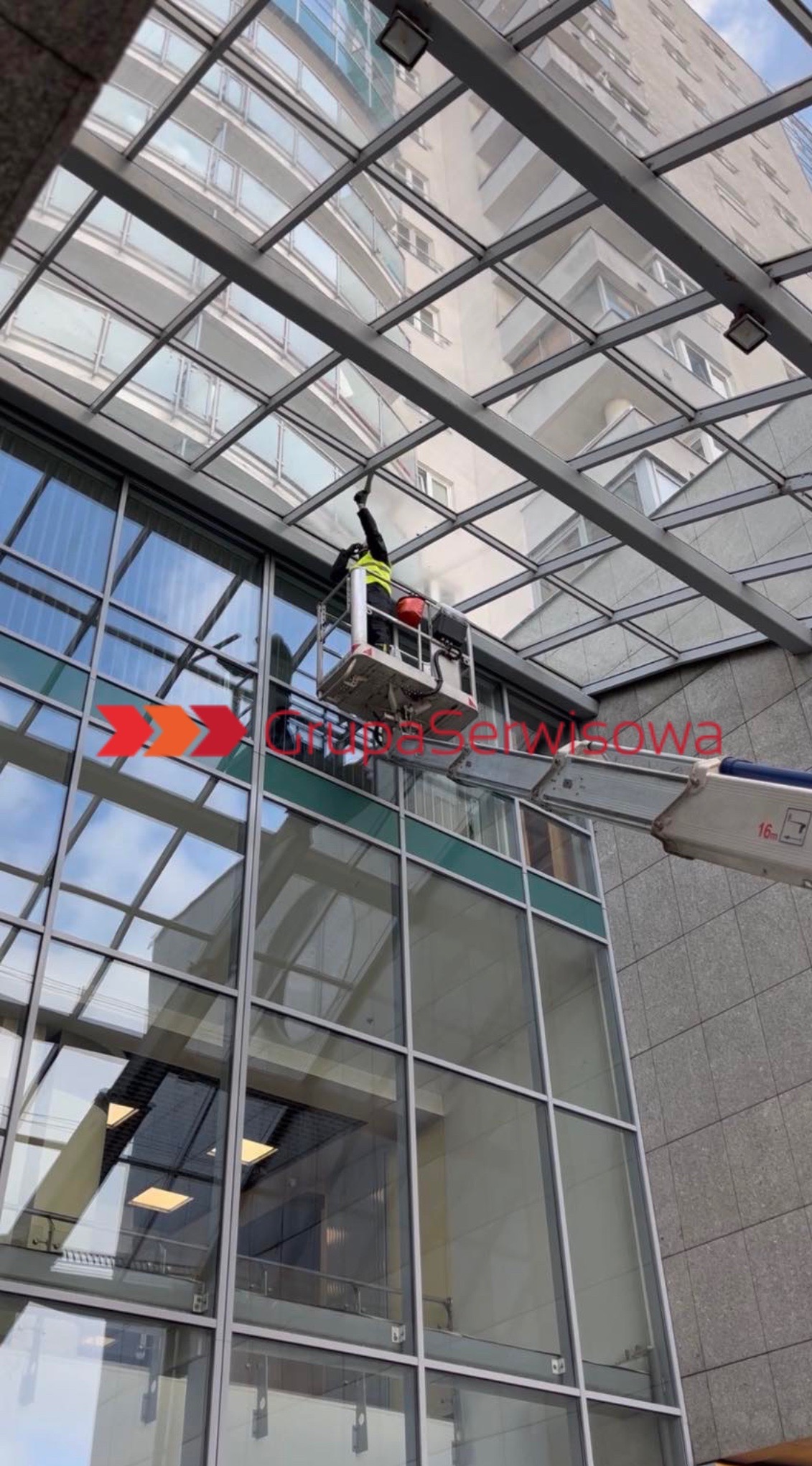 Pracownik w podnośniku czyści szklany dach budynku biurowego. Widoczne odbicia w oknach i konstrukcja dachu. Ujęcie z dołu, z perspektywy przechodnia.