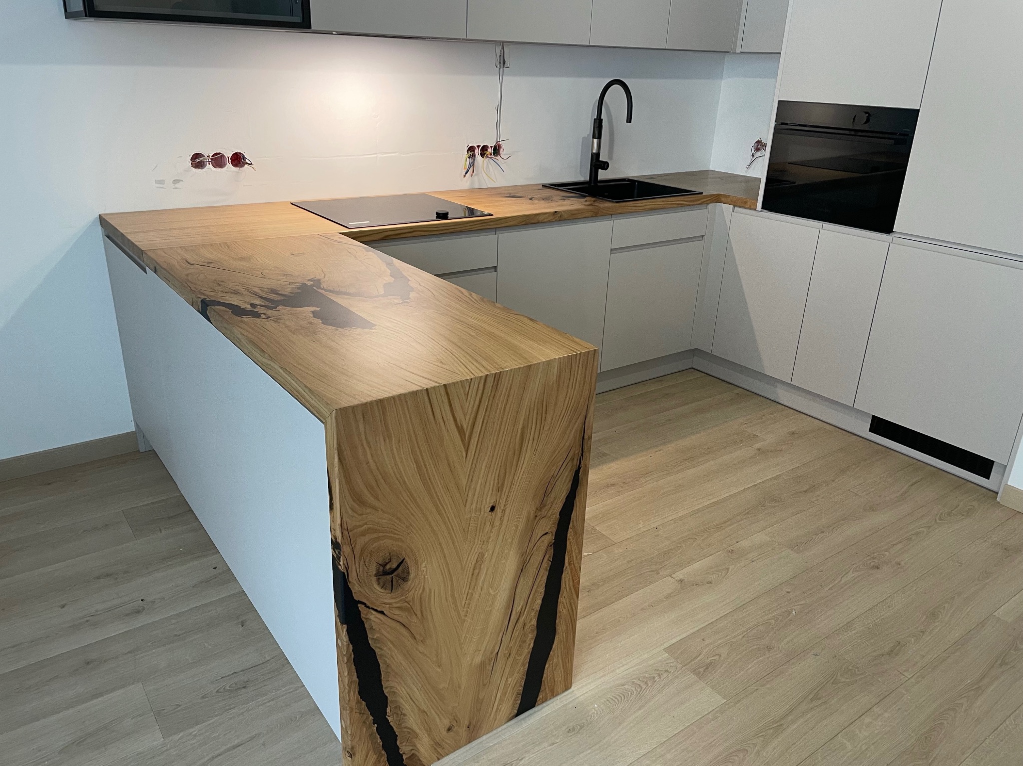 Nowoczesna kuchnia z blatem drewnianym z żywicą epoksydową, czarna armatura i płyta indukcyjna, widoczne niedokończone przyłącza elektryczne.