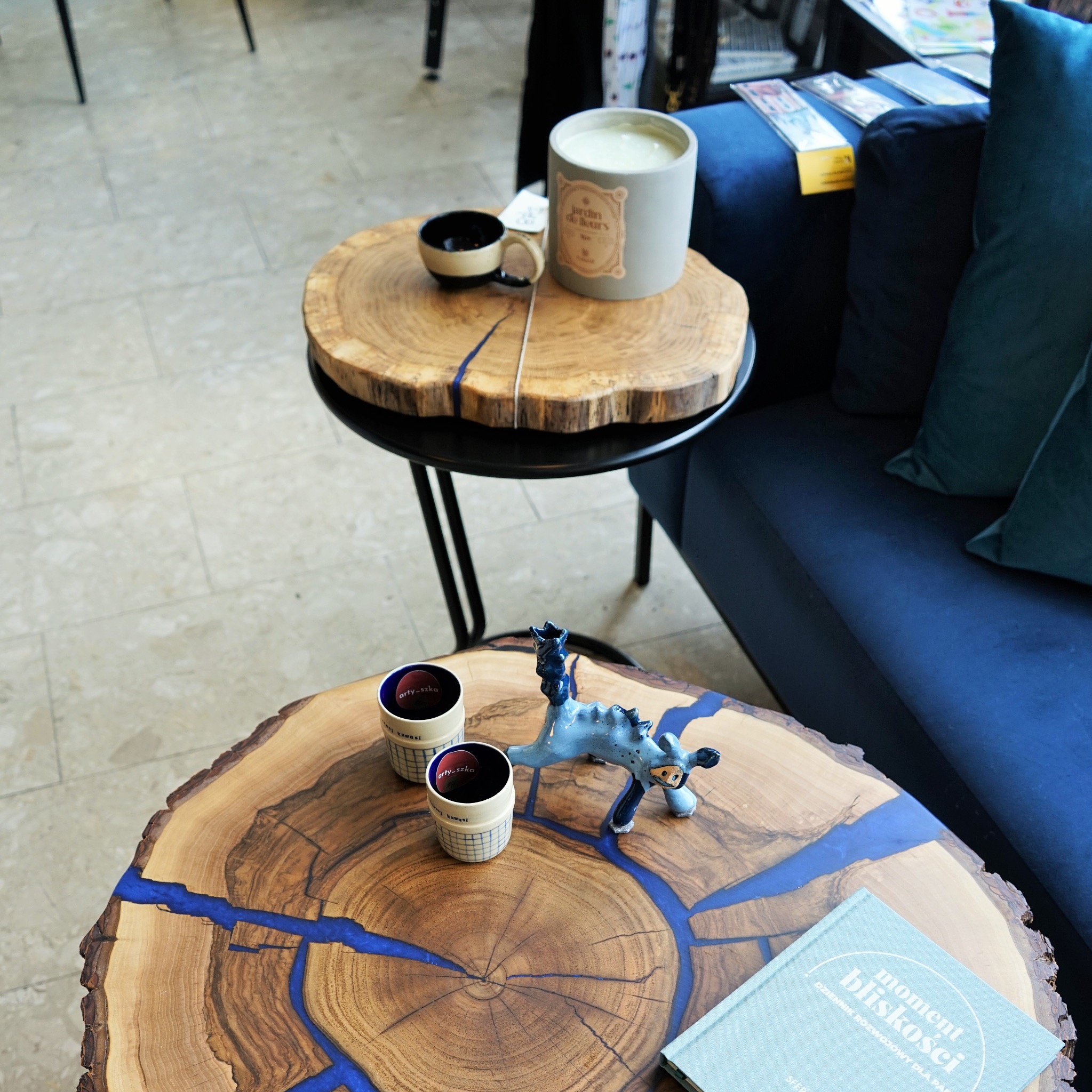 Dwa stoliki kawowe z blatami z plastra drewna wypełnionego niebieską żywicą epoksydową, na jednym filiżanki i ceramiczna figurka, na drugim świeca i filiżanka, obok granatowa sofa.