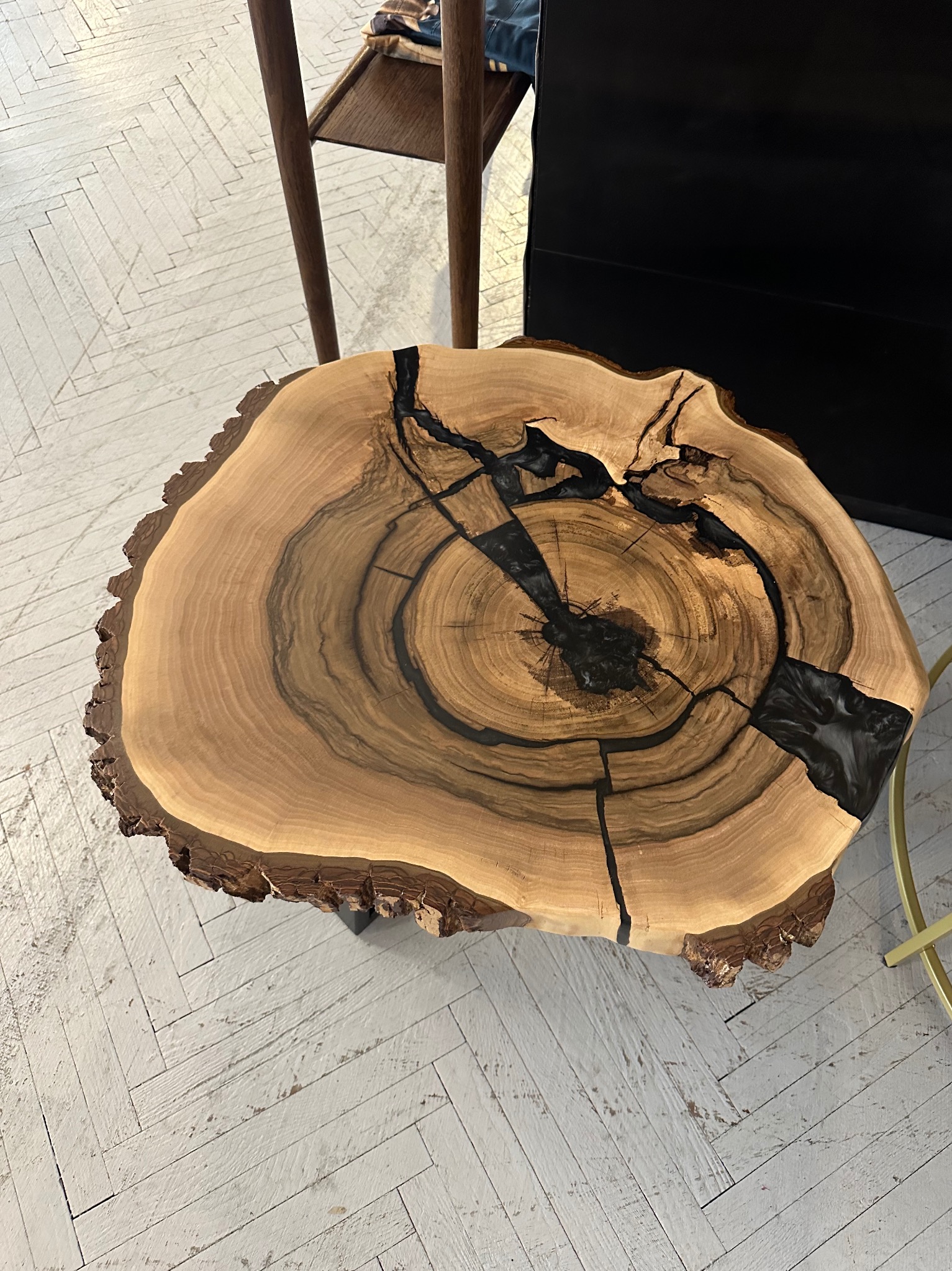 Blat stolika kawowego z plastra drewna z wypełnieniem z czarnej żywicy epoksydowej, widoczne słoje i naturalna krawędź, na tle białej podłogi w jodełkę.