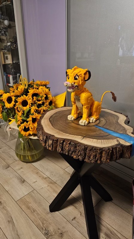 Stolik kawowy z blatem z plastra drewna z żywicą epoksydową, na którym stoi figurka lwa z klocków, obok bukiet słoneczników z dekoracją '40'.