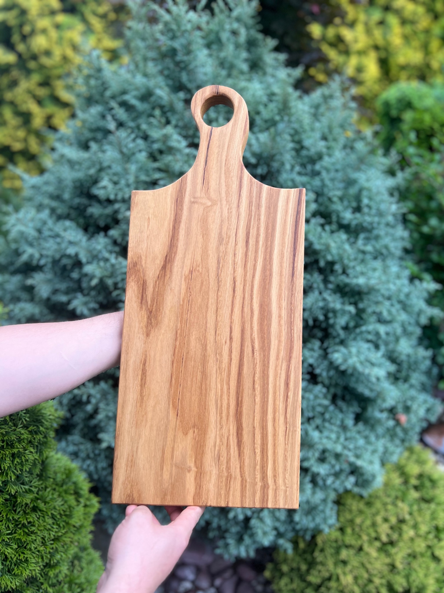 Drewniana deska do krojenia z uchwytem, prezentowana na tle zielonych krzewów